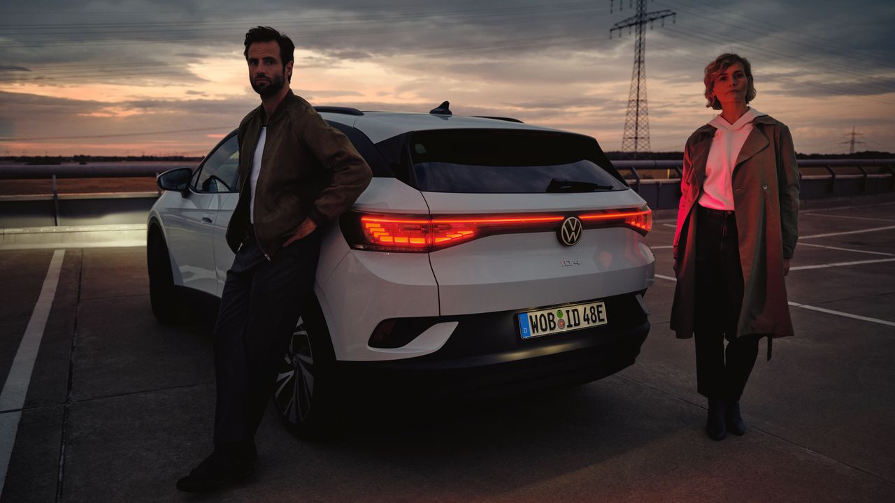 VW ID.4 branco parado no estacionamento no escuro. Um homem e uma mulher encontram-se junto à traseira.