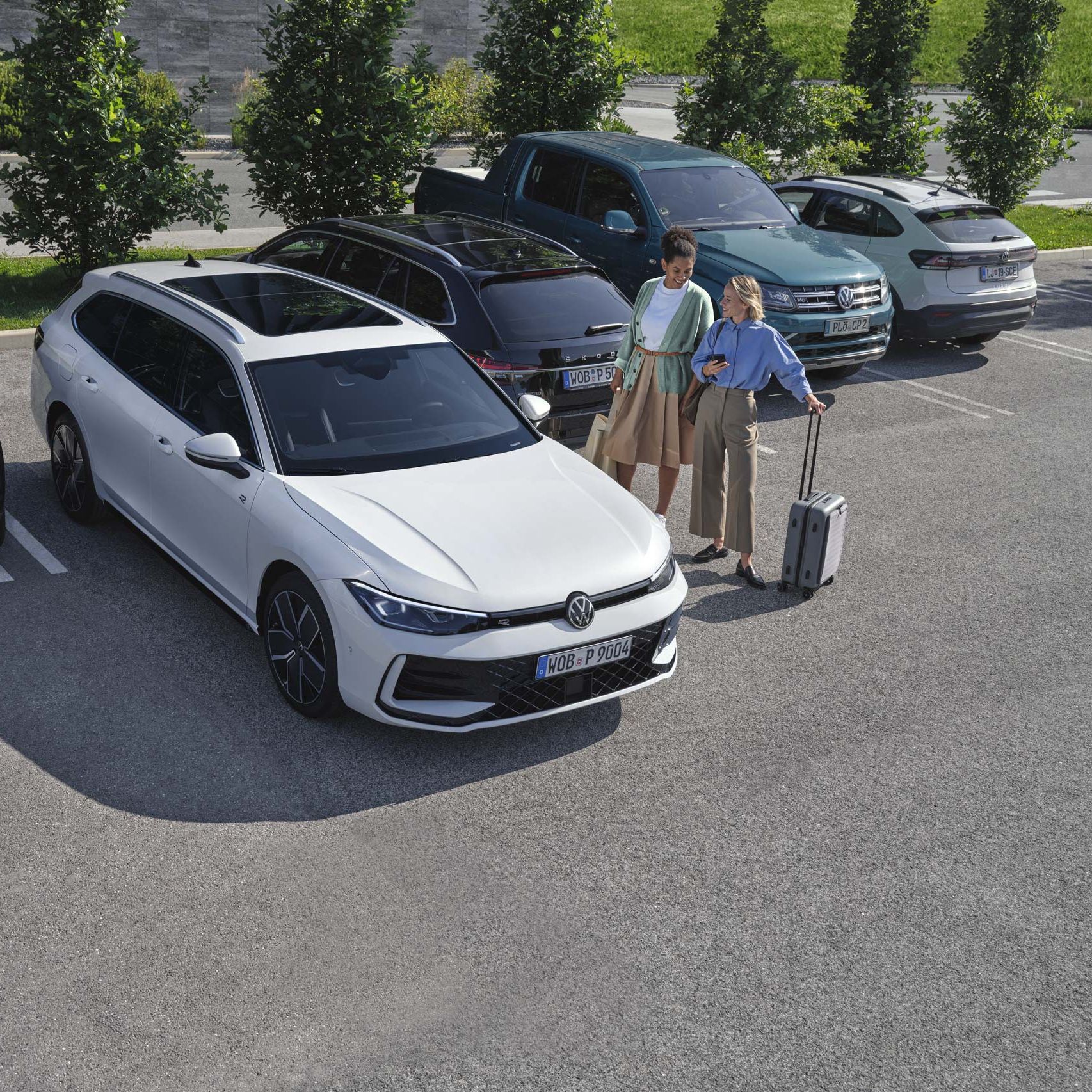 Duas mulheres do lado de fora do VW Passat branco, que estaciona por elas utilizando o sistema opcional Park Assist Plus. Uma mulher segura o seu smartphone na mão.