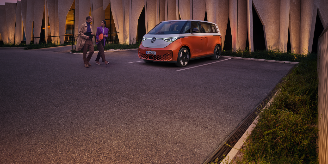 Ein weiß-gelber VW ID. Buzz steht auf einem Platz, umgeben von einer Familie. Front und Seite des Fahrzeugs sichtbar.