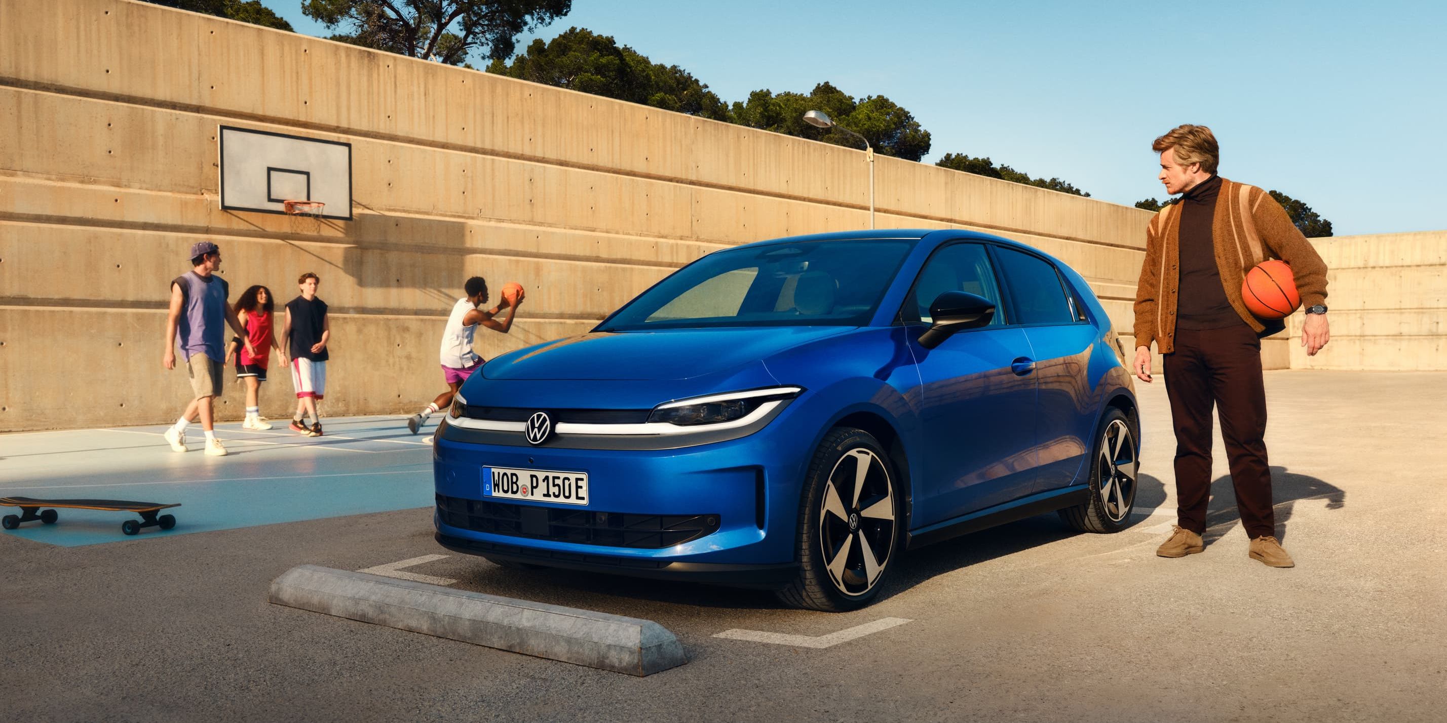 Um VW ID. Polo azul estacionado junto a um campo de basquete. Um homem está de pé nas proximidades, segura uma bola de basquetebol debaixo do braço.