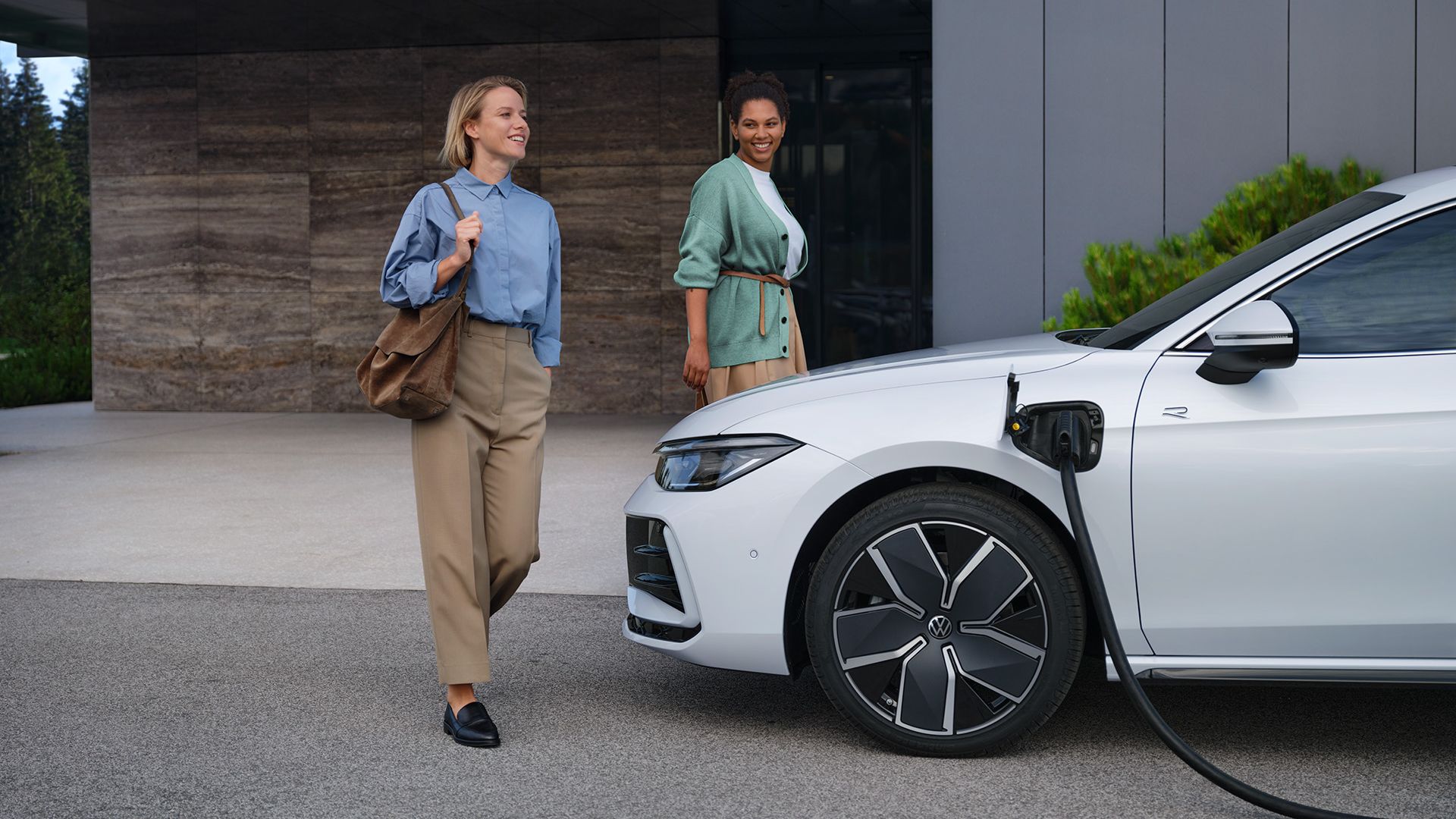 VW Passat branco lateralmente a um edifício de escritórios com o cabo de carregamento ligado, duas mulheres aproximam-se do carro.