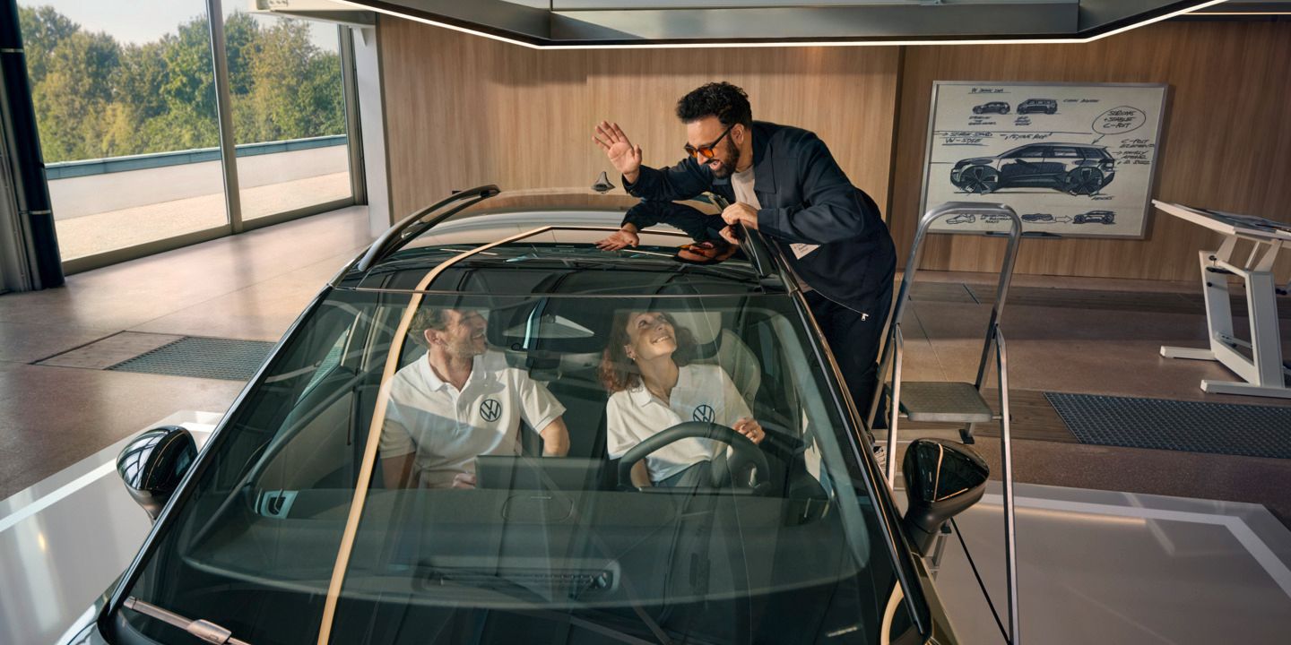 Um homem encontra-se numa escada ao lado de um VW ID. Cross a acenar pelo teto panorâmico às pessoas sentadas no seu interior.