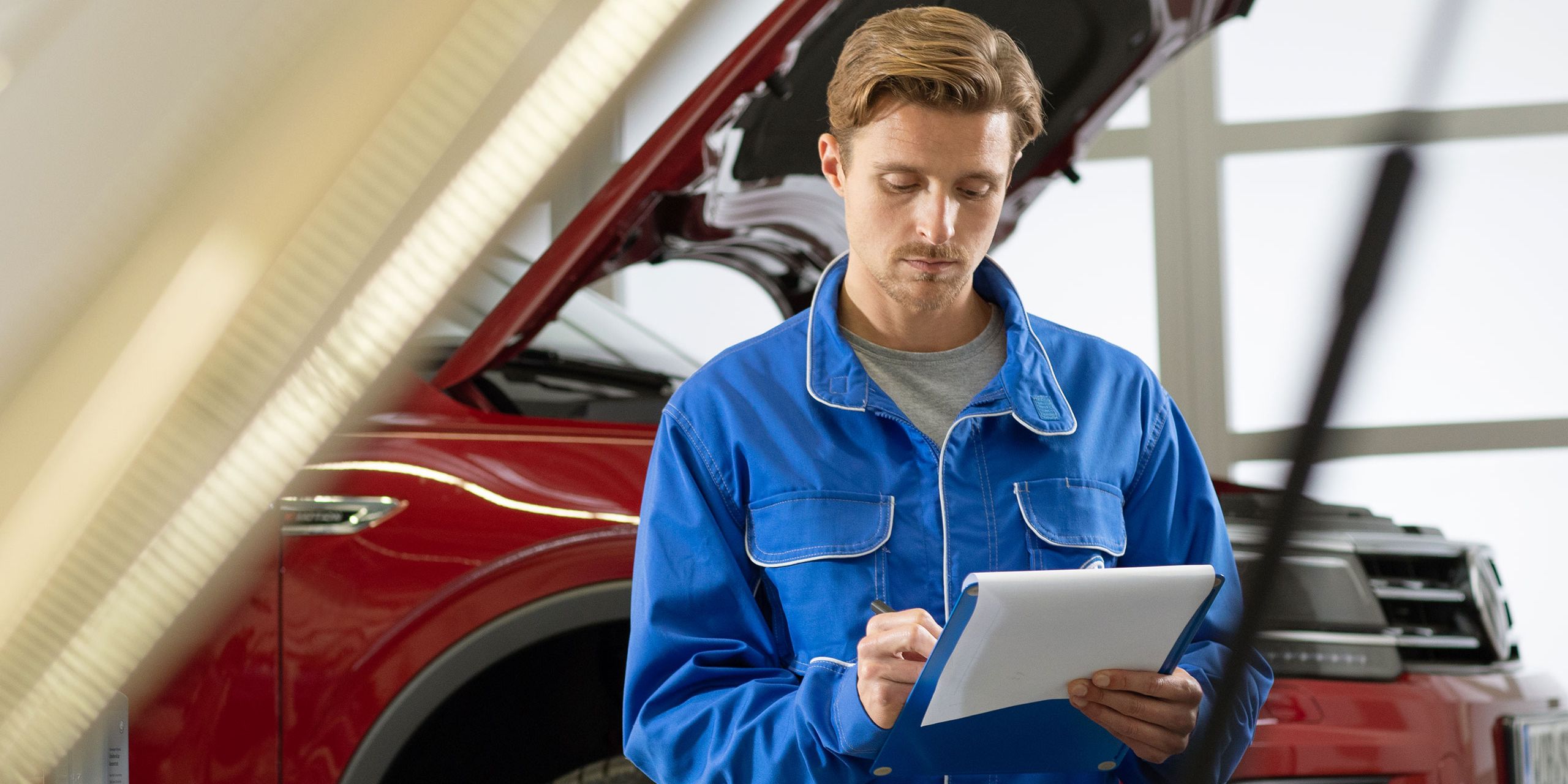 Funcionário do serviço VW verifica uma lista de verificação em frente de um VW vermelho numa rampa hidráulica com capô aberto