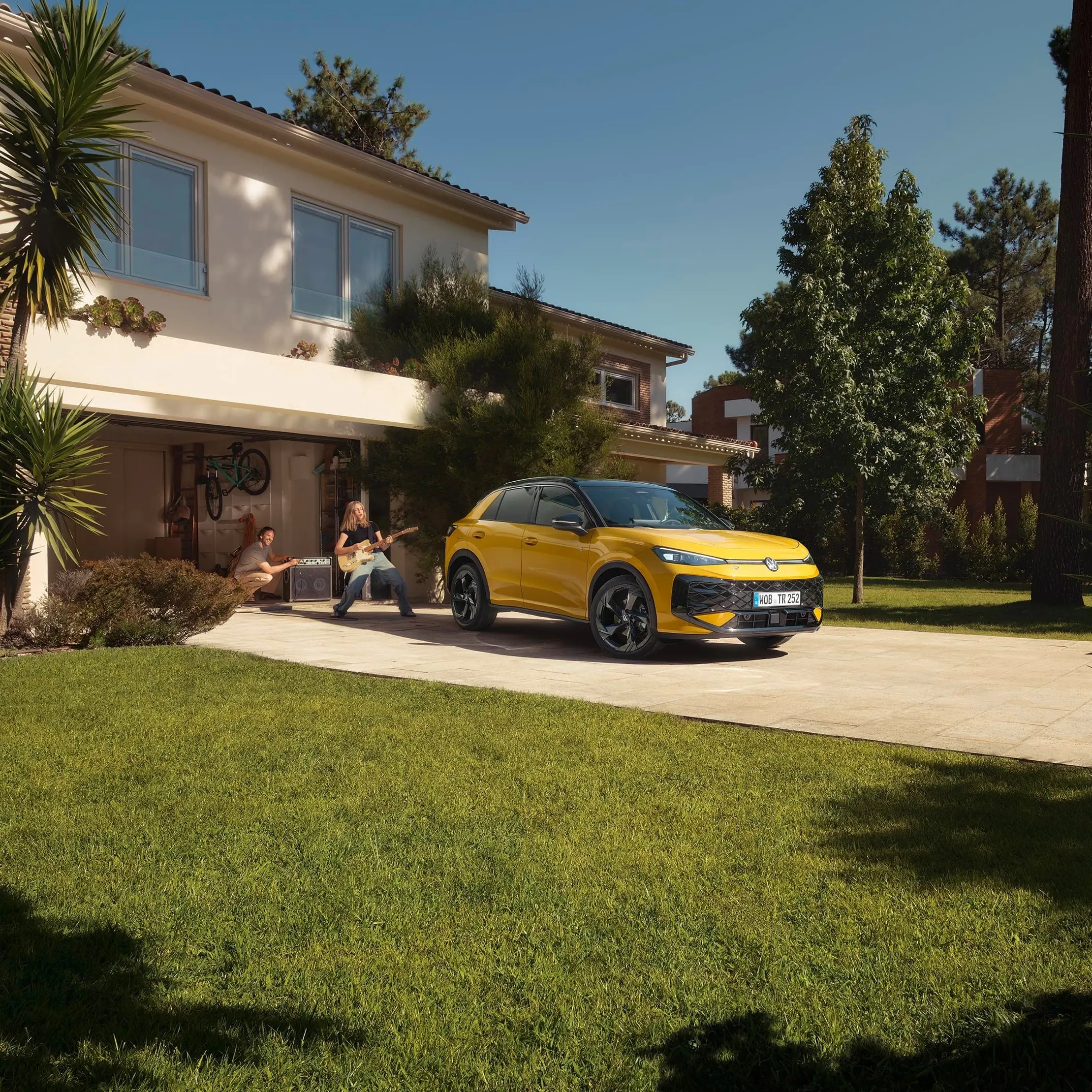 Um VW T-Roc R-Line amarelo em frente à garagem, uma jovem a tocar guitarra com o pai a controlar o altifalante.