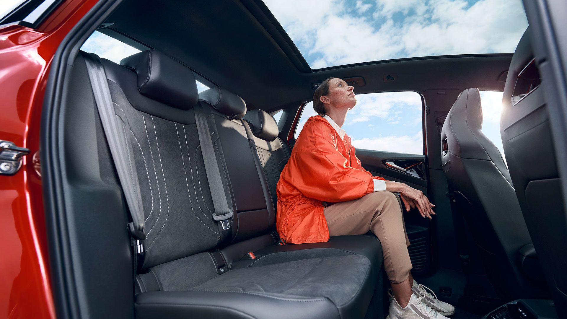Uma mulher jovem sentada no banco traseiro de um VW ID.5 GTX vermelho a olhar pelo teto panorâmico para o céu azul.
