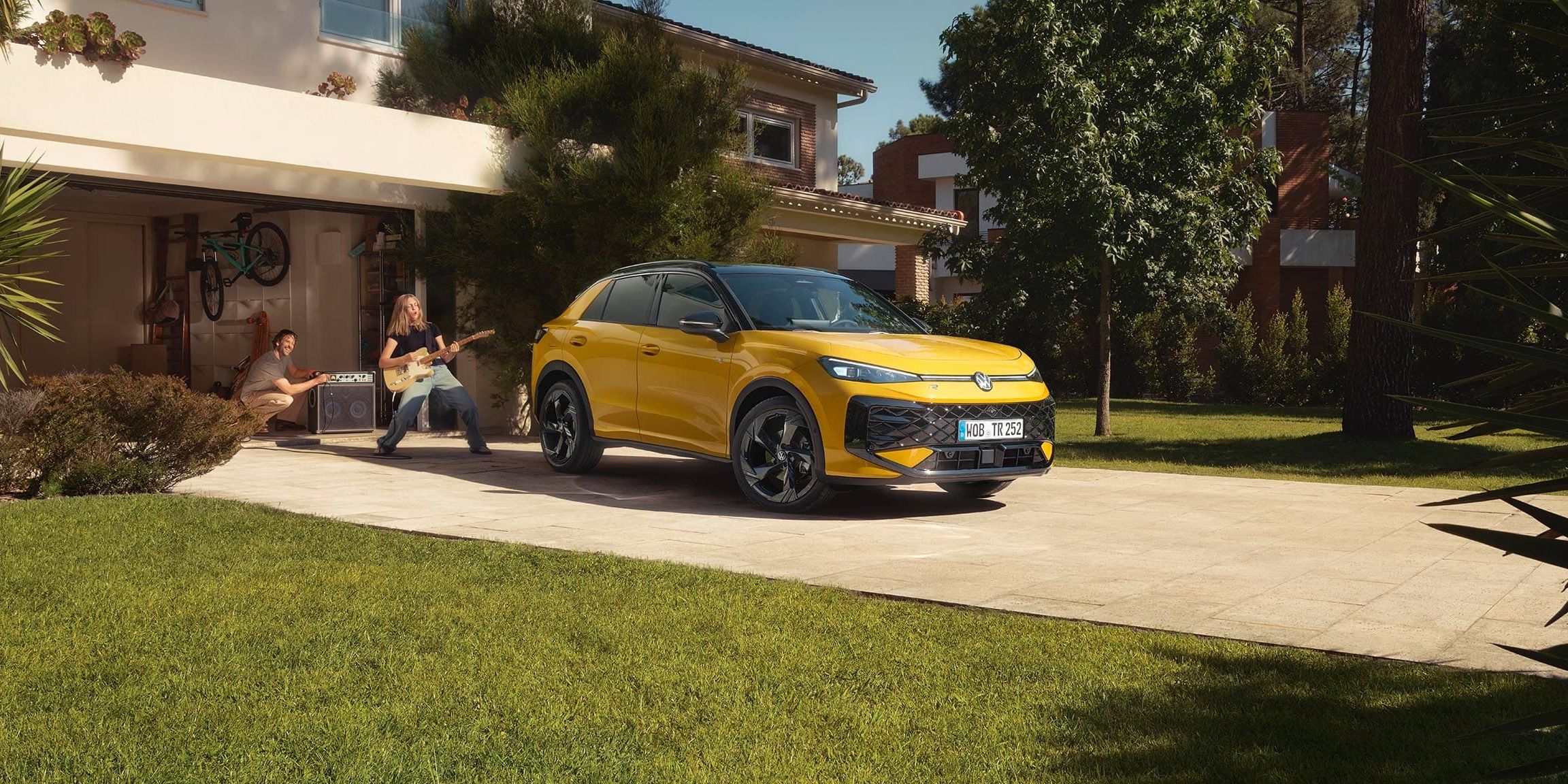 Um VW T-Roc R-Line amarelo em frente à garagem, uma jovem a tocar guitarra com o pai a controlar o altifalante.