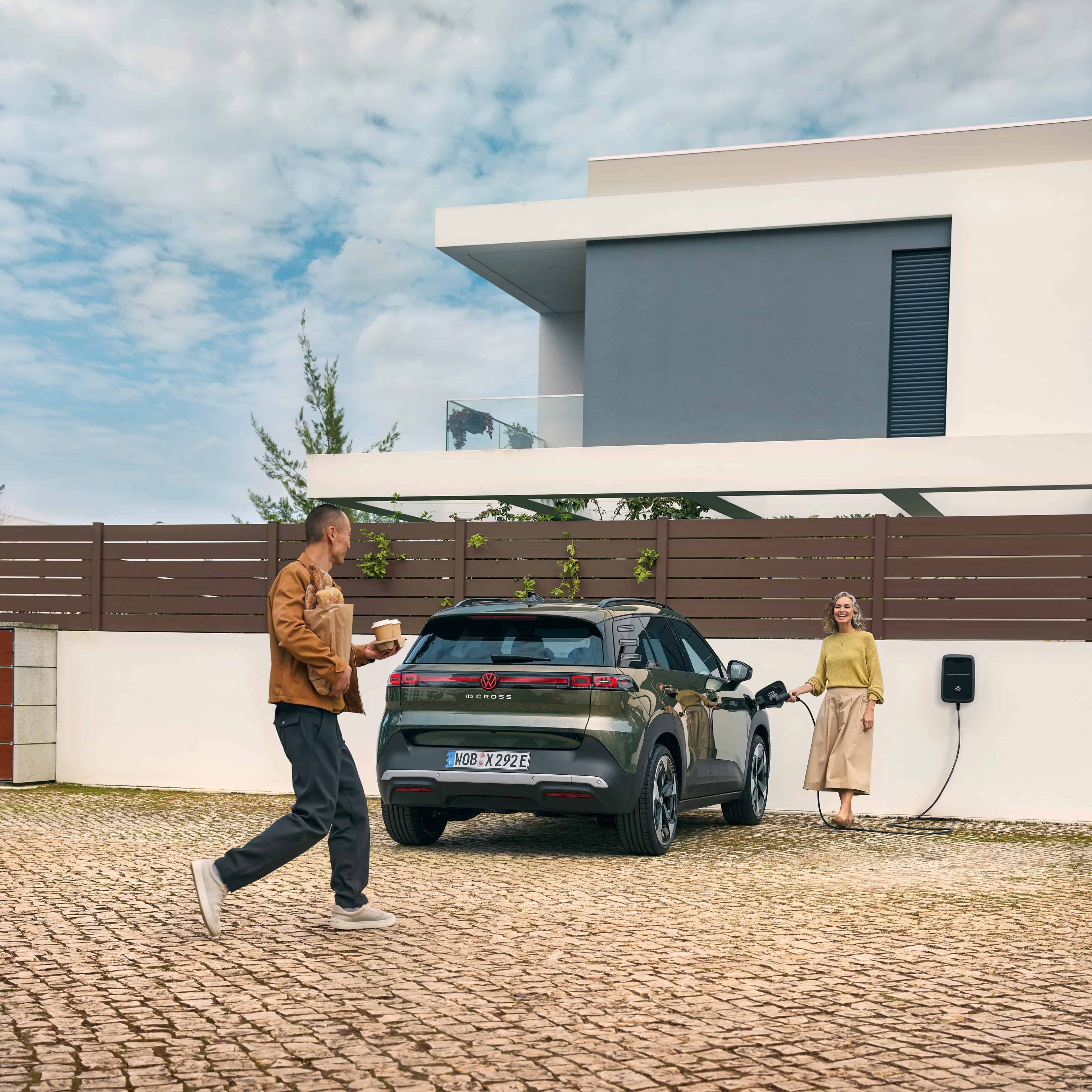 Homem caminha em direção a uma casa moderna enquanto segura um copo de café e pão. Mulher a inserir o carregador da wallbox de casa no VW ID. Cross   