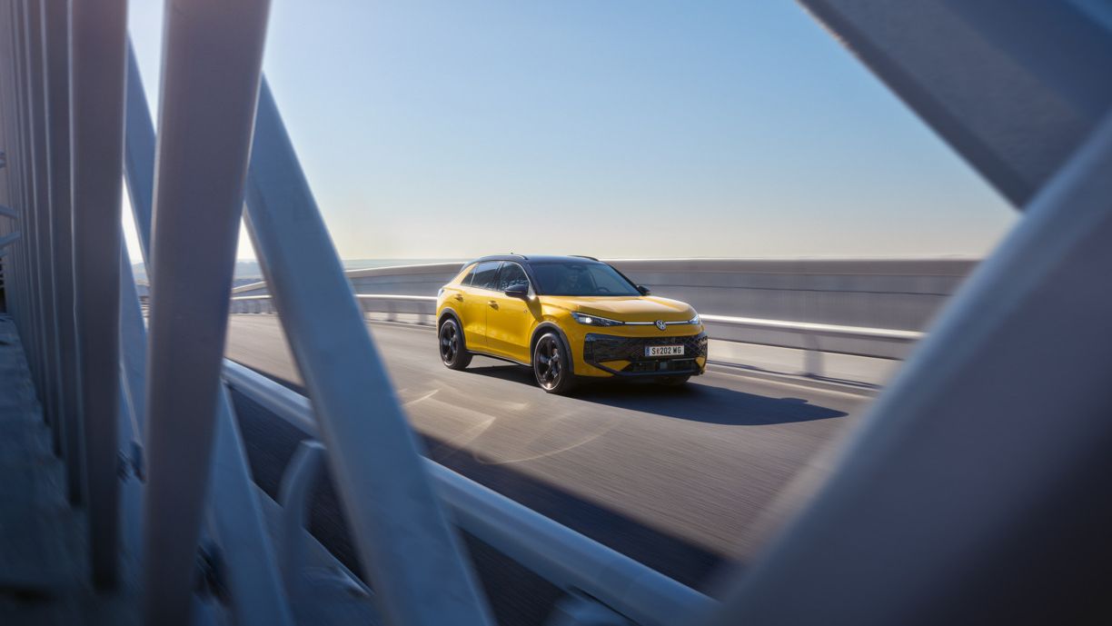 Um VW T-Roc R-Line amarelo circula numa ponte com uma estrutura em aço moderna.