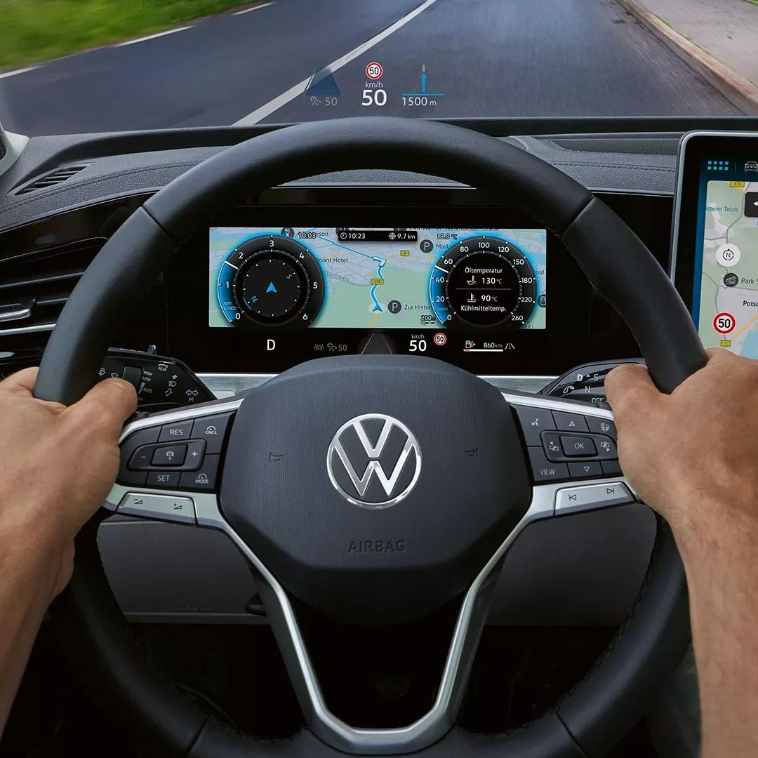 Vista interior do VW Passat focada no lado do condutor com um grande ecrã a cores e o head-up display. Duas mãos agarram o volante. 