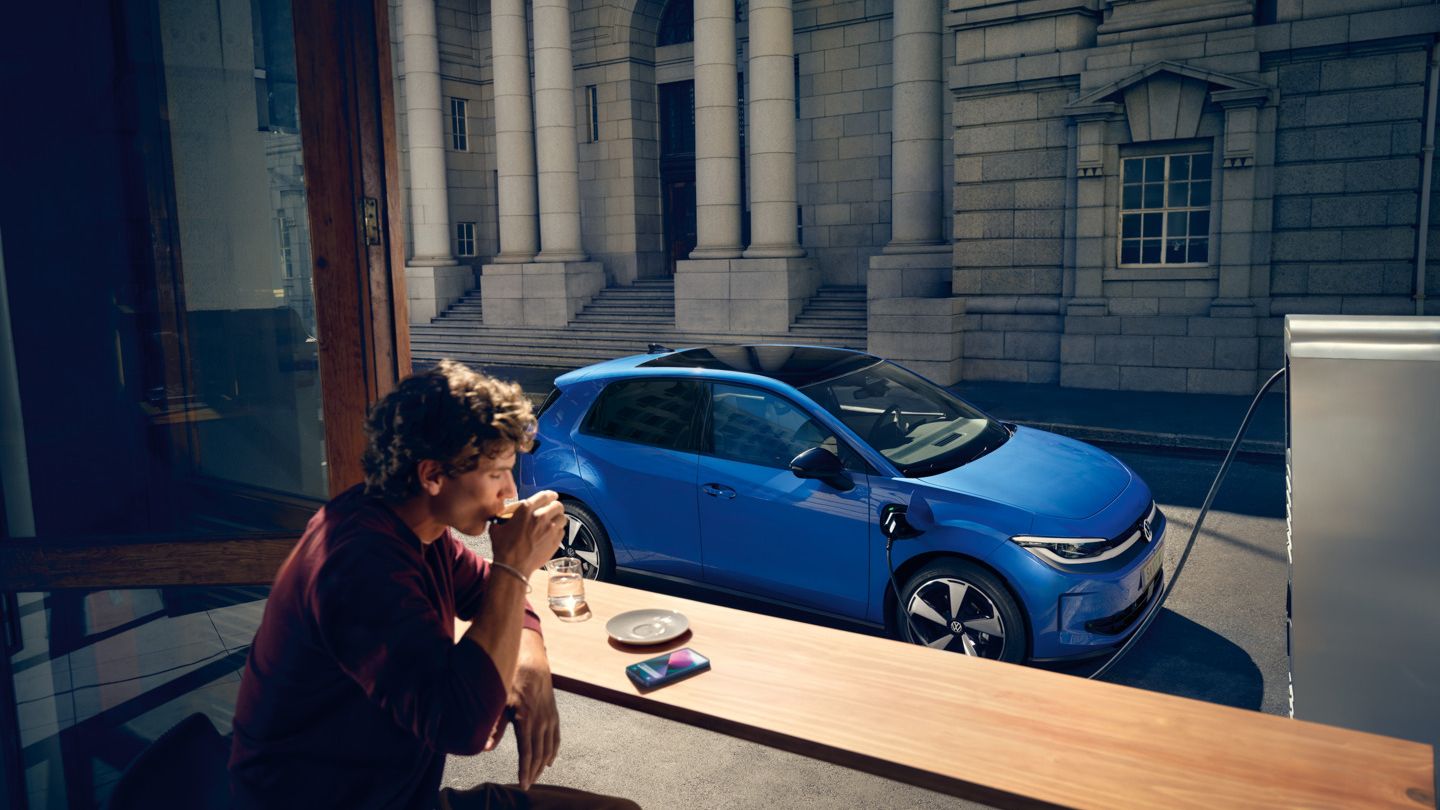 Um homem está sentado num café, através de uma janela aberta vê-se o VW ID. Polo a carregar numa estação de carregamento.
