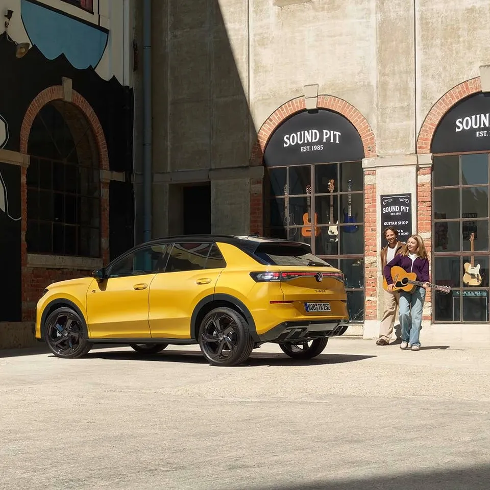 Pai e filha com uma guitarra junto de um VW T-Roc R-Line amarelo estacionado em frente a uma loja de música.