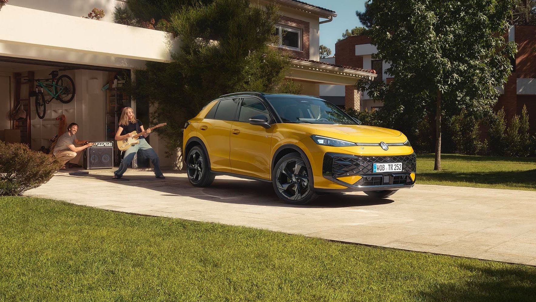 Um VW T-Roc R-Line amarelo em frente à garagem, uma jovem a tocar guitarra com o pai a controlar o altifalante.