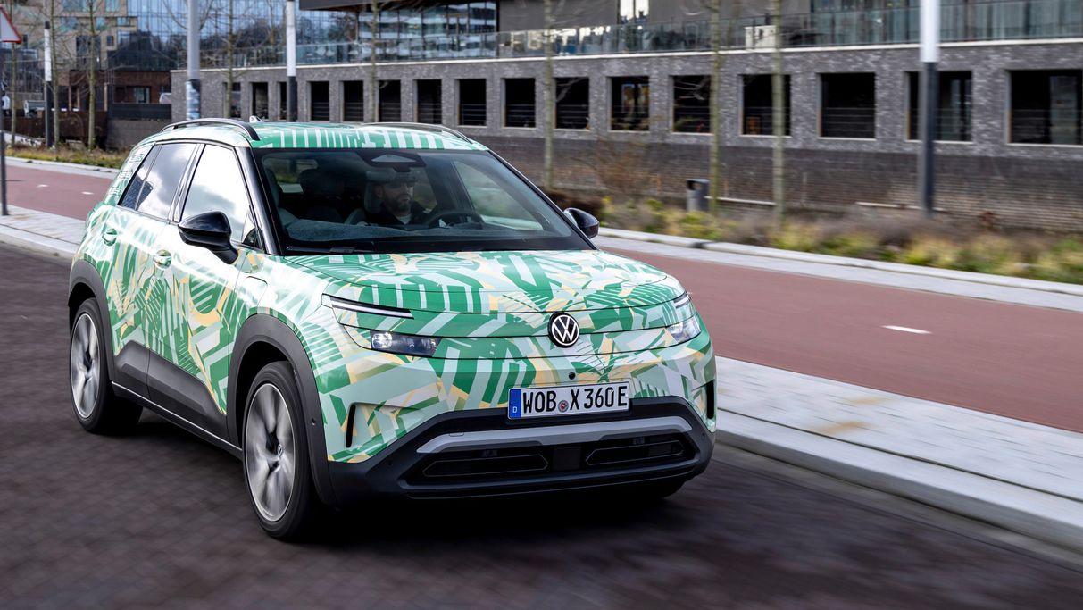 Ein Elektroauto fährt auf einer städtischen Straße an modernen Gebäuden vorbei. Das Fahrzeug hat ein auffälliges Design mit grünen und gelben Mustern. Im Hintergrund ist ein Gebäude mit der Aufschrift "Mitsubishi Electric Football Club" zu sehen.