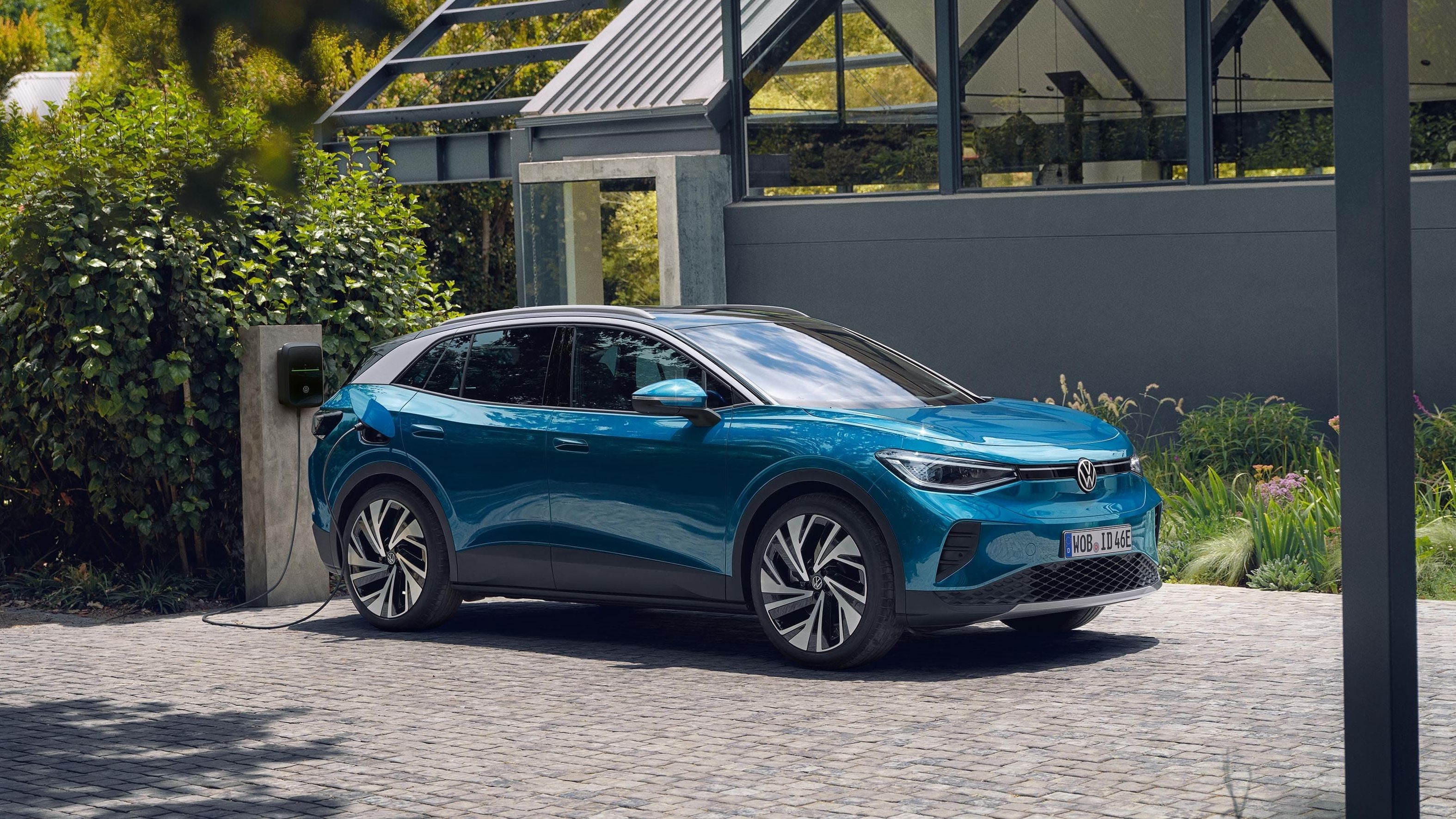 Um VW ID.4 azul estacionado lateralmente em frente de uma casa moderna e a carregar numa Wallbox.