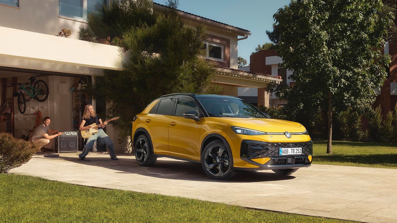 Um VW T-Roc R-Line amarelo em frente à garagem, uma jovem a tocar guitarra com o pai a controlar o altifalante.