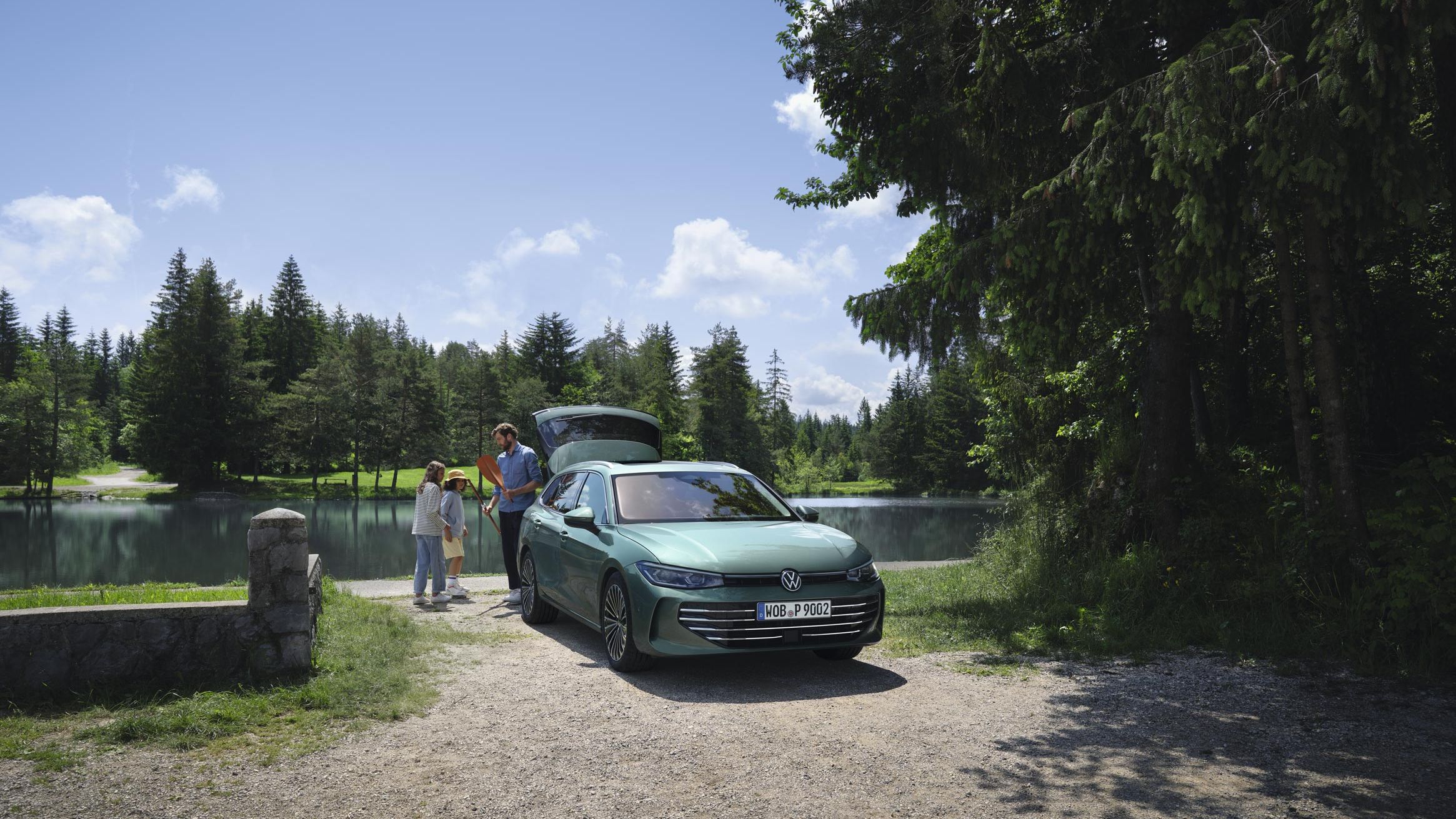 VW Passat verde-azulado estacionado num lago com a bagageira aberta. Um homem distribui brinquedos de lazer a duas crianças.