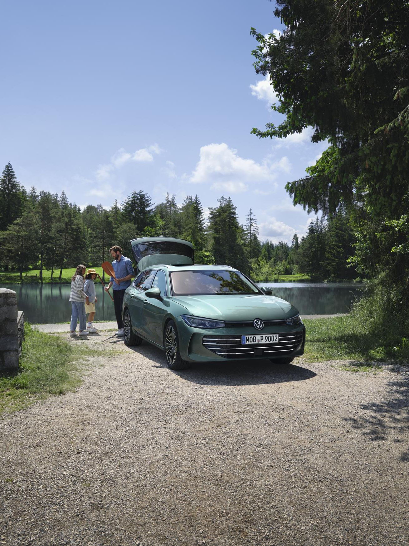 VW Passat verde-azulado estacionado num lago com a bagageira aberta. Um homem distribui brinquedos de lazer a duas crianças.