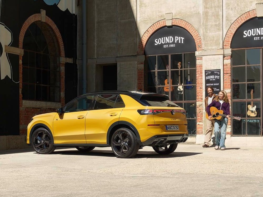 Pai e filha com uma guitarra junto de um VW T-Roc R-Line amarelo estacionado em frente a uma loja de música.