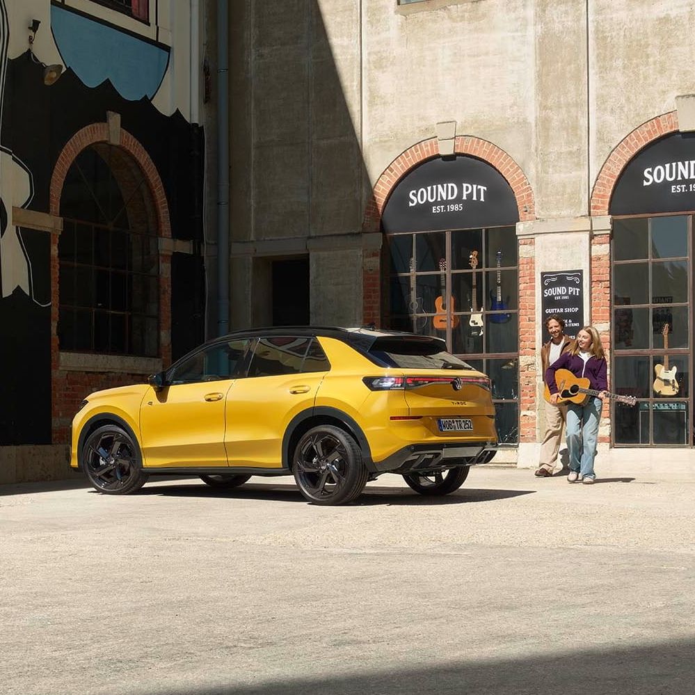 Pai e filha com uma guitarra junto de um VW T-Roc R-Line amarelo estacionado em frente a uma loja de música.