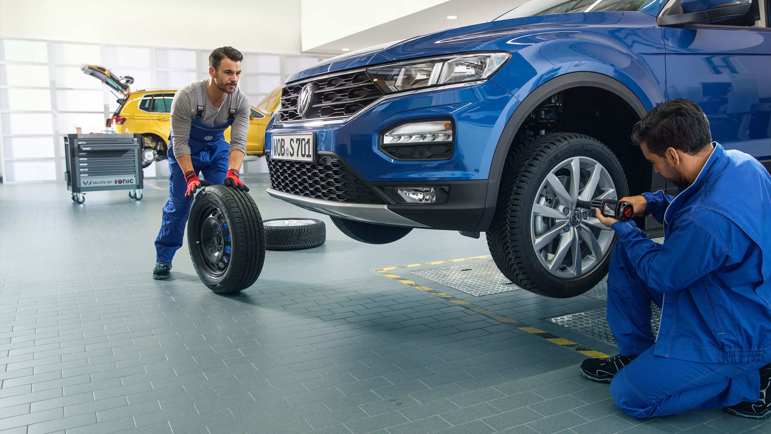 Dois empregados do serviço VW trabalham juntos na troca de pneus de um VW Tiguan azul numa oficina