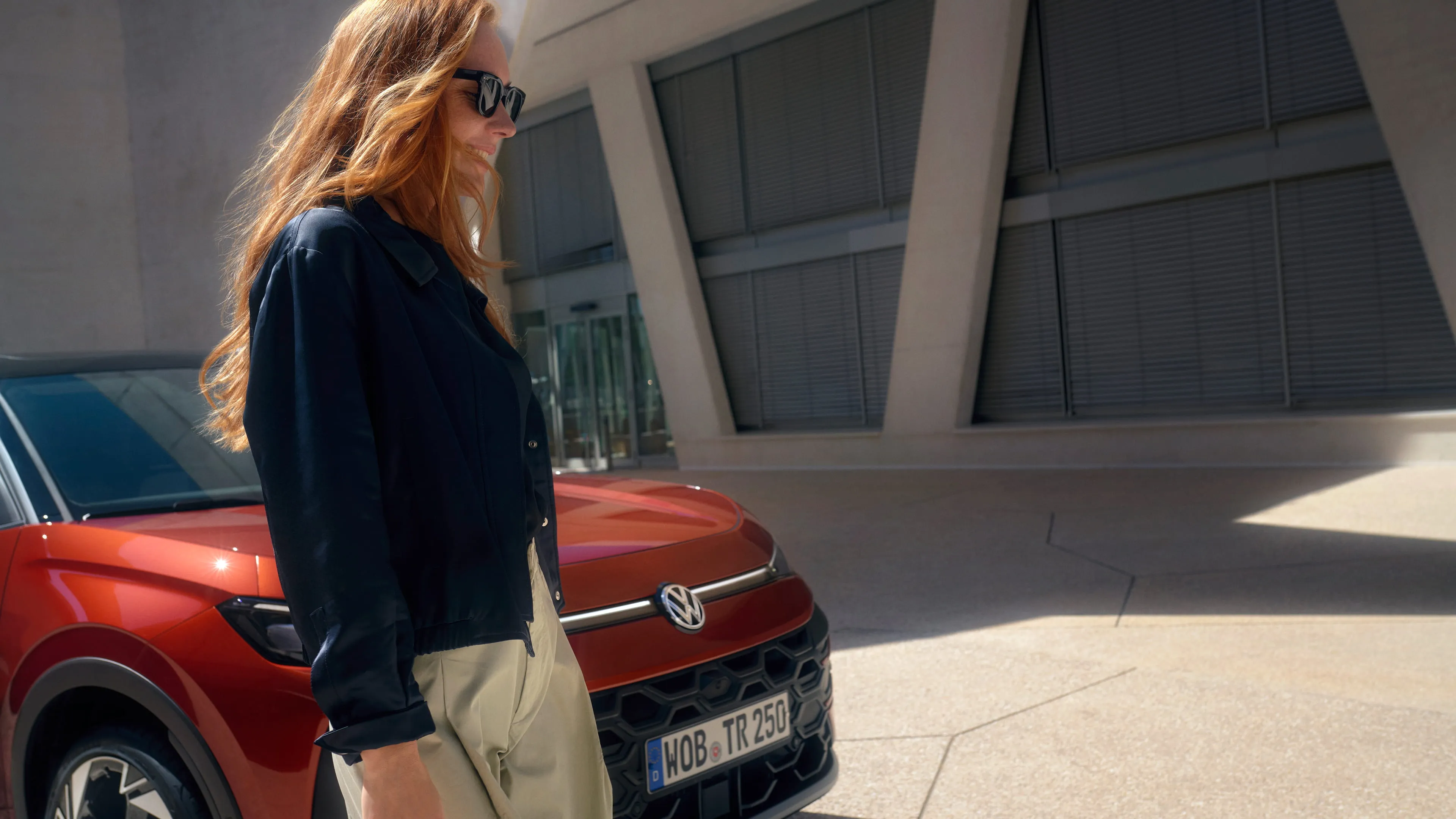 Uma mulher com óculos de sol junto a um VW T-Roc Style vermelho, em frente a um edifício.