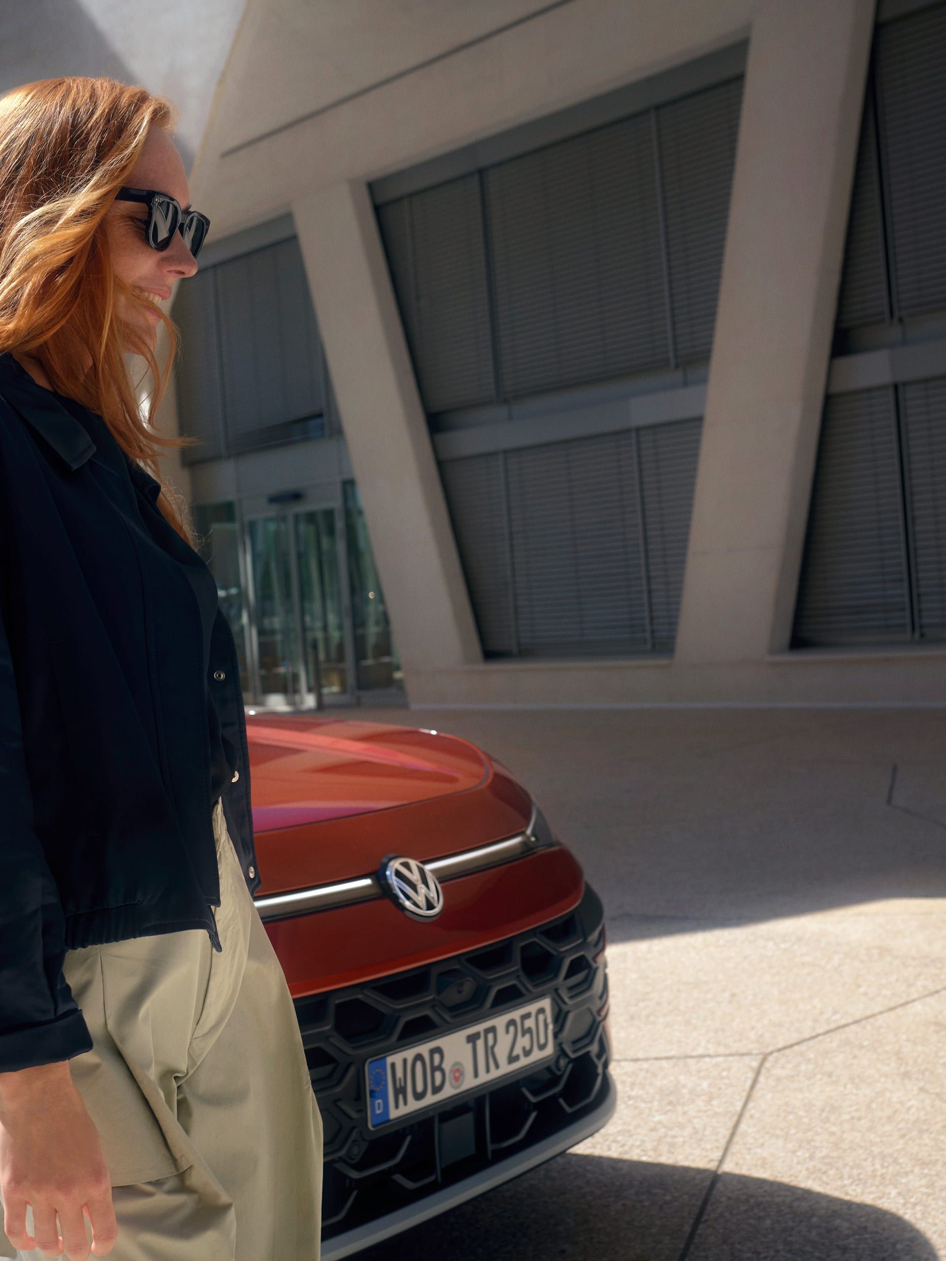 Uma mulher com óculos de sol junto a um VW T-Roc Style vermelho, em frente a um edifício.