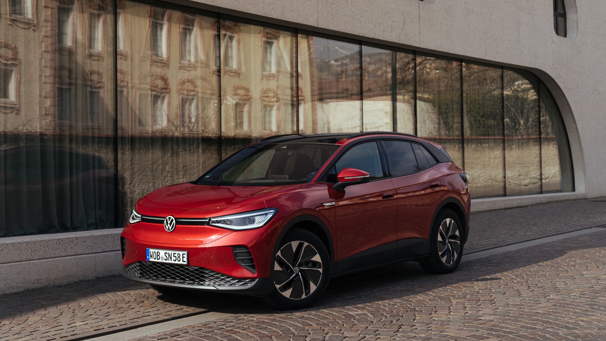 Veículo elétrico Volkswagen ID.4 de cor vermelha estacionado em frente a um edifício moderno com janelas altas e estreitas.