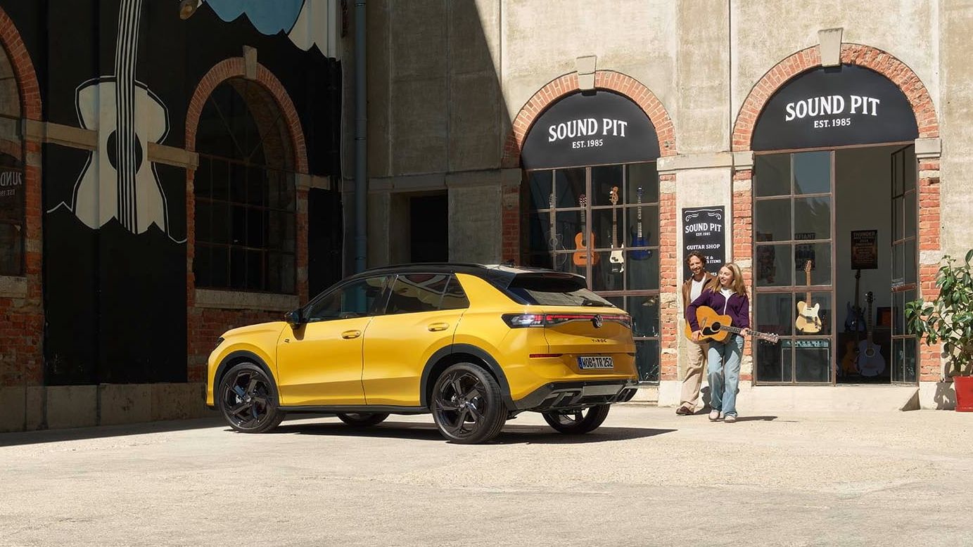 Pai e filha com uma guitarra junto de um VW T-Roc R-Line amarelo estacionado em frente a uma loja de música.
