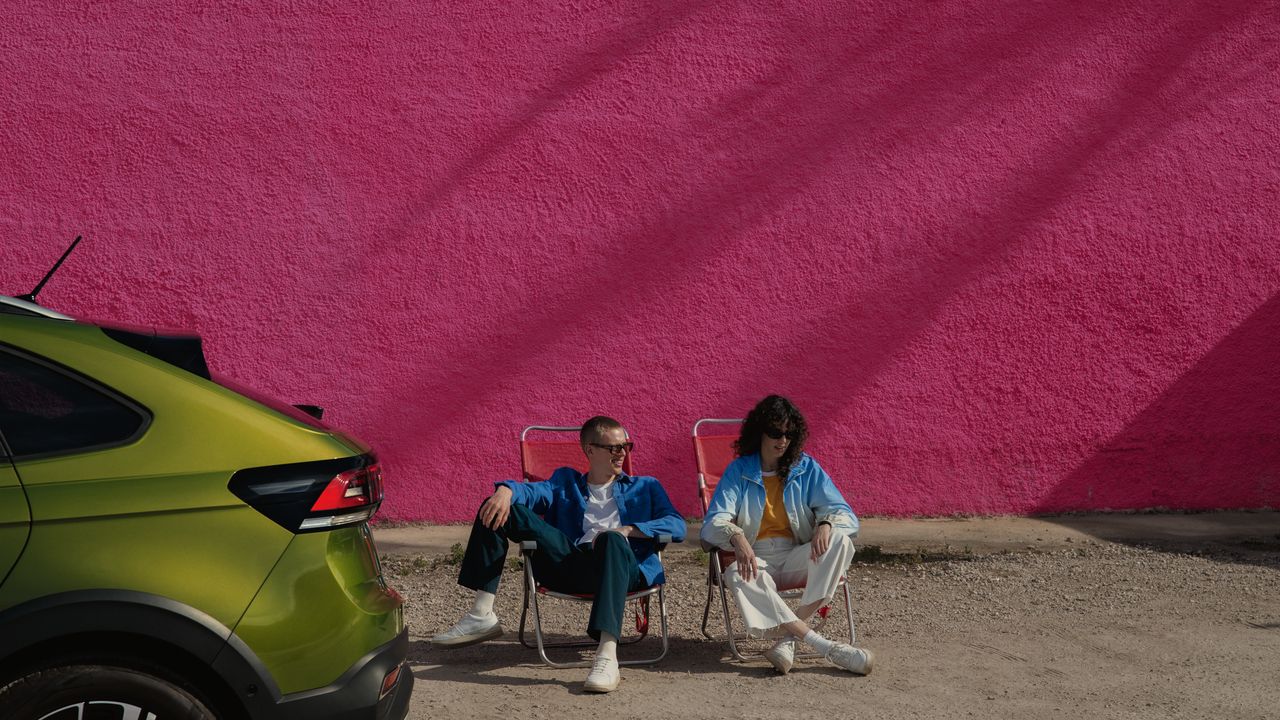 Traseira de VW Taigo verde; atrás duas pessoas sentadas em cadeiras de campismo
