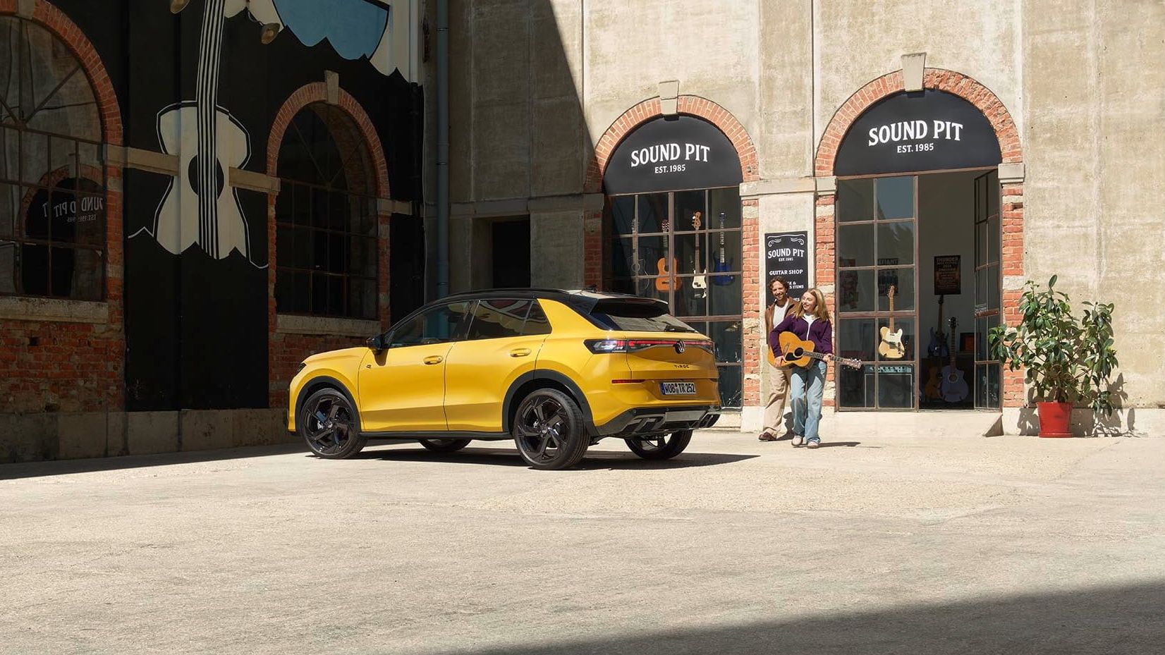 Pai e filha com uma guitarra junto de um VW T-Roc R-Line amarelo estacionado em frente a uma loja de música.