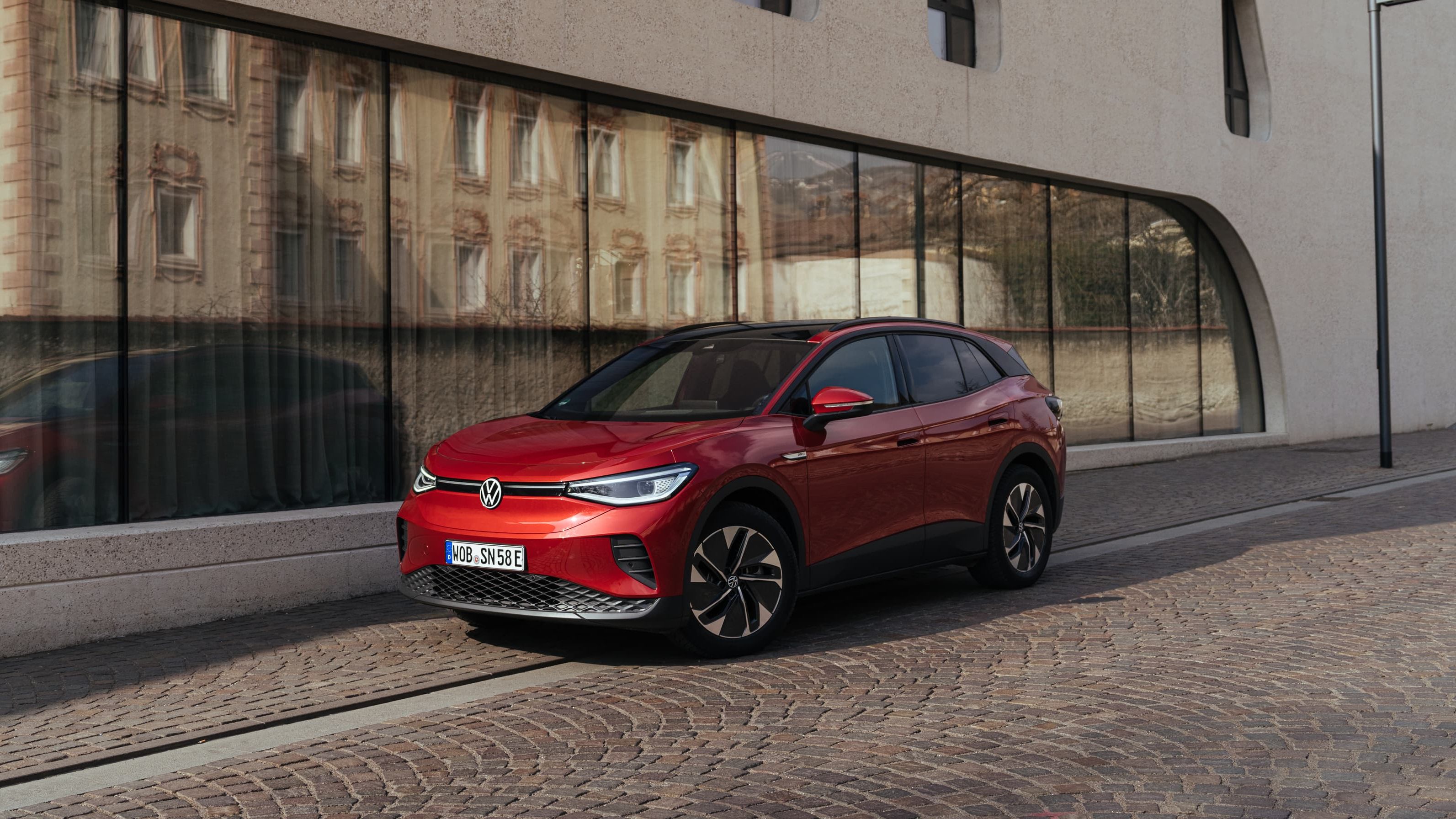 Veículo elétrico Volkswagen ID.4 de cor vermelha estacionado em frente a um edifício moderno com janelas altas e estreitas.