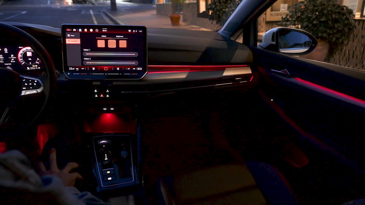 Das Bild zeigt das Innere eines Autos bei Nacht. Die Beleuchtung ist rot und eine Hand ist in der Nähe des Schalthebels sichtbar. Ein großer Bildschirm mit einem Interface ist auf dem Armaturenbrett zu sehen.