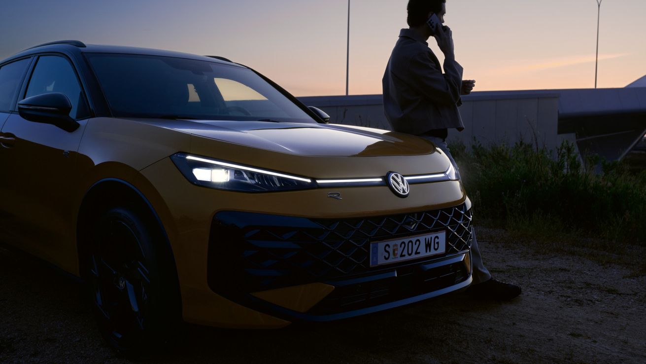 Um homem junto a um VW T-Roc R-Line com os faróis ligados e o logótipo iluminado.