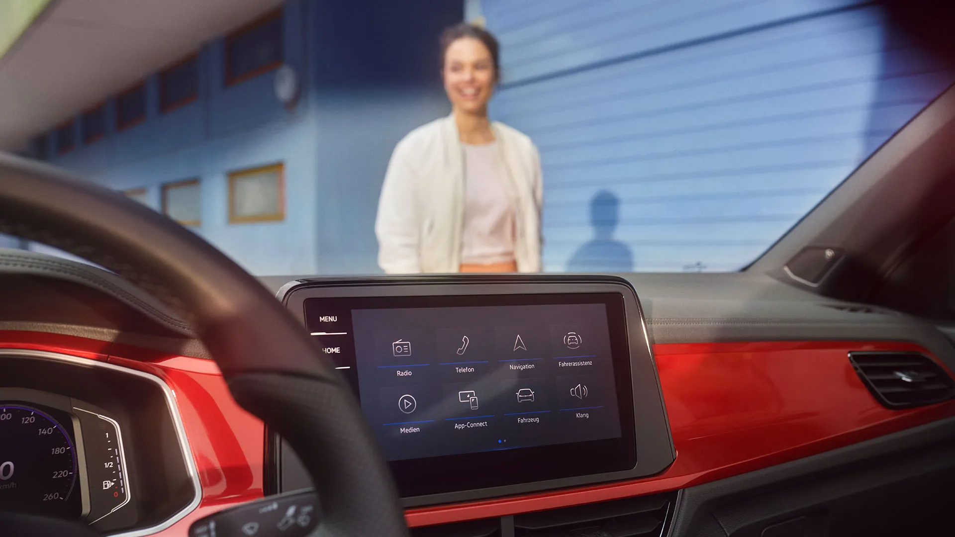 Interior do T-Roc Cabrio: Vista do ecrã com a exibição do menu e o volante multifunções. Uma mulher está de pé, à frente do Cabrio.