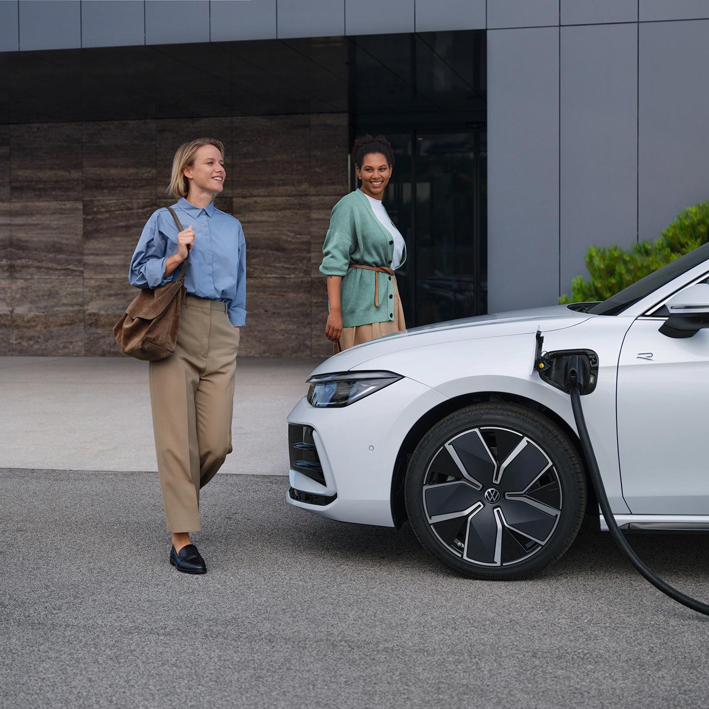 VW Passat branco lateralmente a um edifício de escritórios com o cabo de carregamento ligado, duas mulheres aproximam-se do carro.