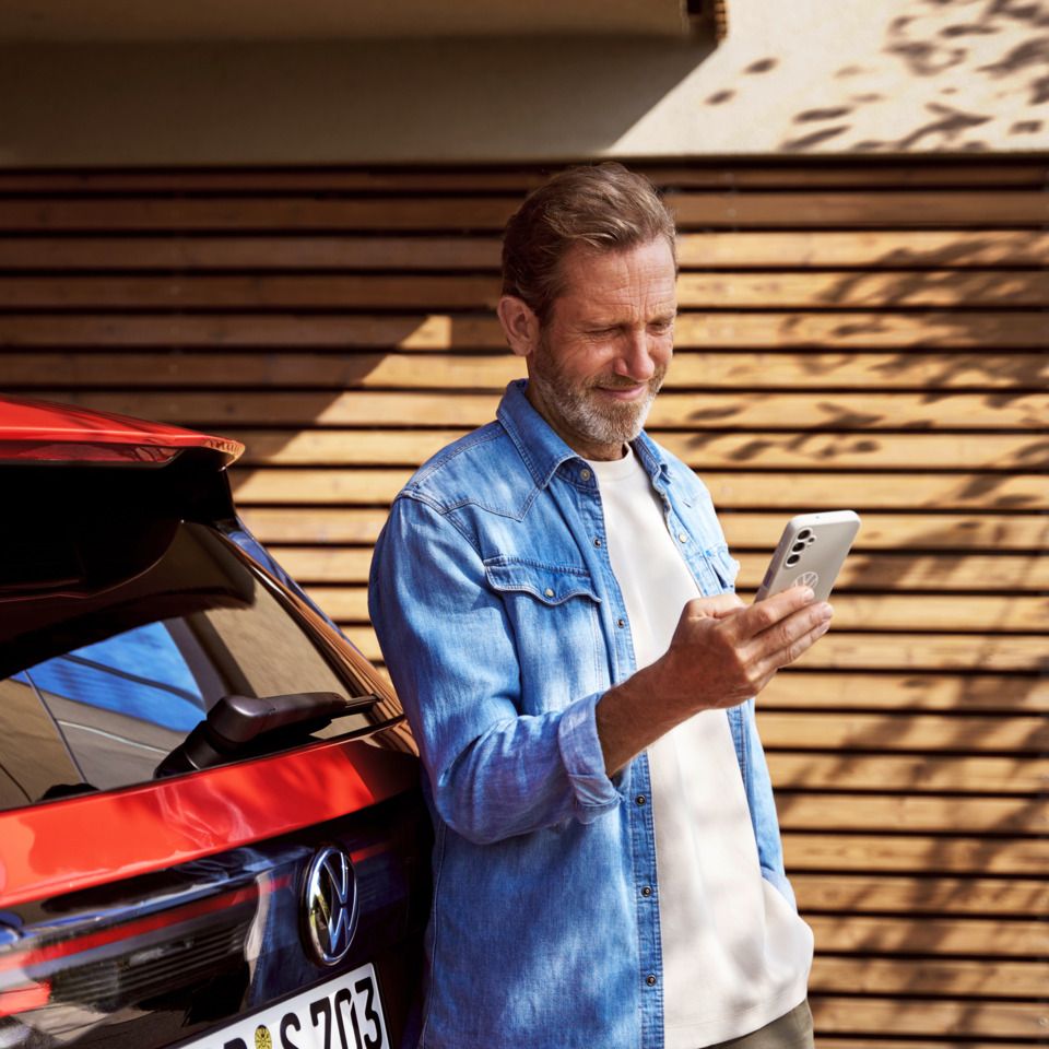Ein Mann mit Smartphone in der Hand steht angelehnt an seinem Tiguan.
