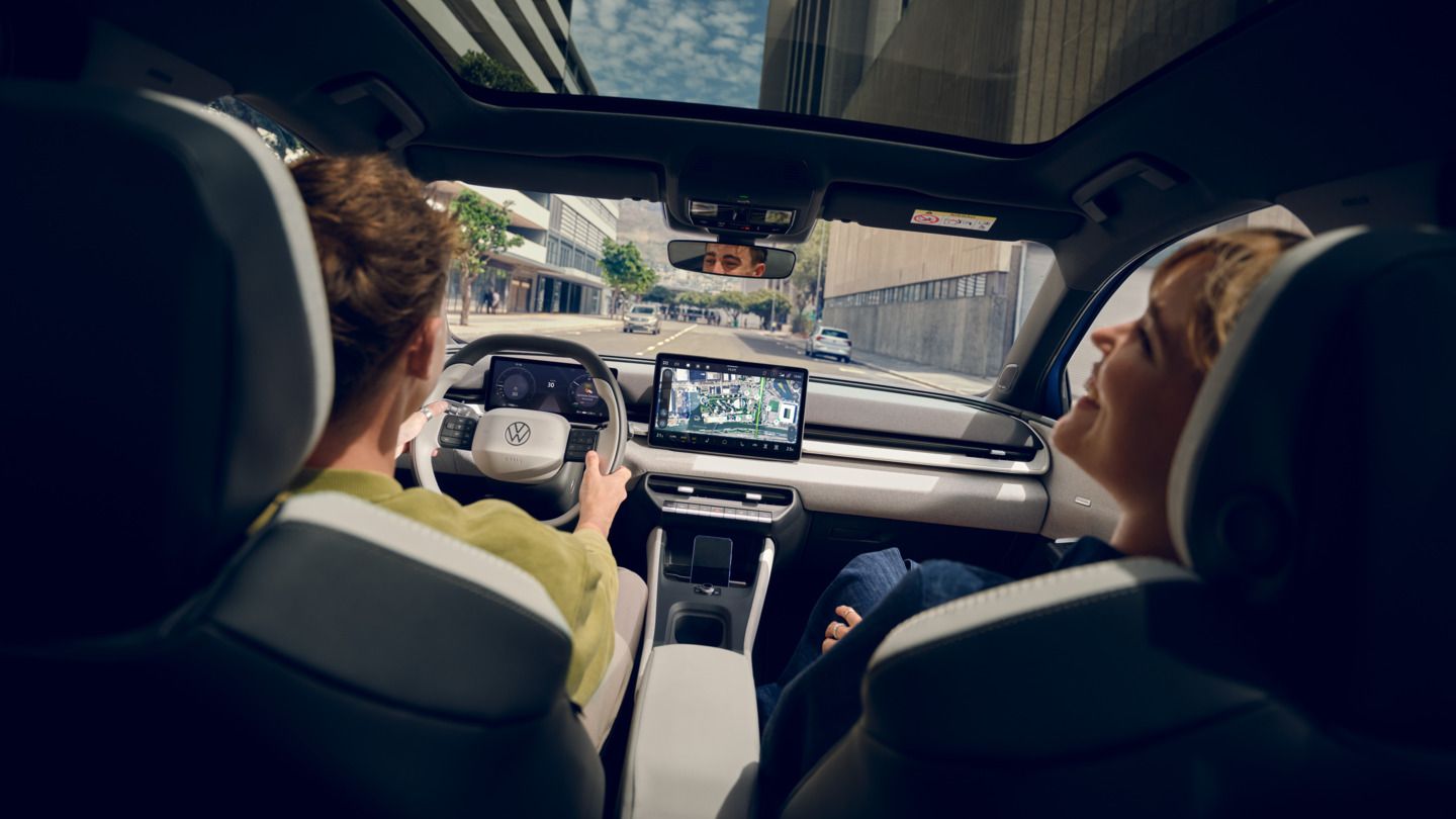 Um homem conduz um Volkswagen ID. Polo, no banco do passageiro uma mulher a rir. Veem-se edifícios altos através do teto de vidro panorâmico.