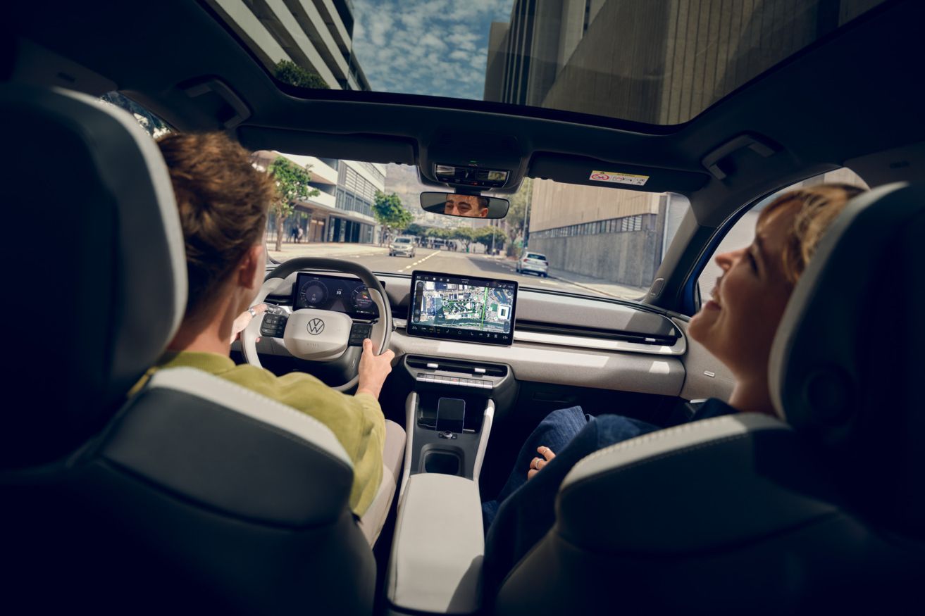 Um homem conduz um Volkswagen ID. Polo, no banco do passageiro uma mulher a rir. Veem-se edifícios altos através do teto de vidro panorâmico.