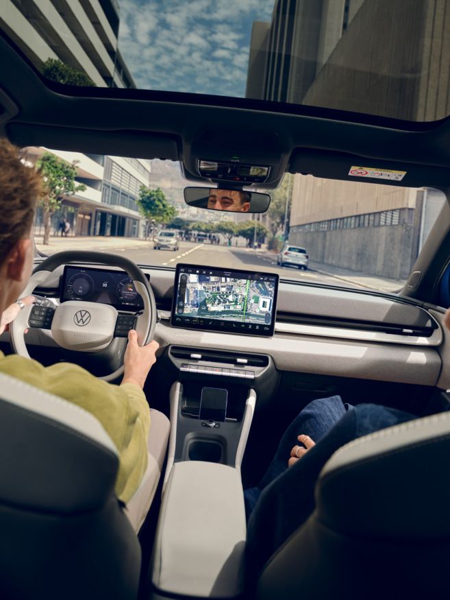 Um homem conduz um Volkswagen ID. Polo, no banco do passageiro uma mulher a rir. Veem-se edifícios altos através do teto de vidro panorâmico.