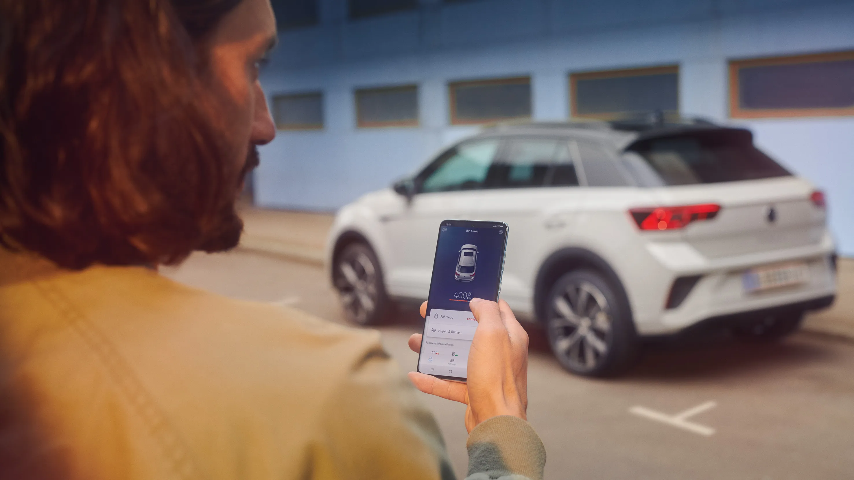 VW T-Roc R-Line em branco em frente de um edifício, vê-se a lateral, um homem em primeiro plano com smartphone na mão