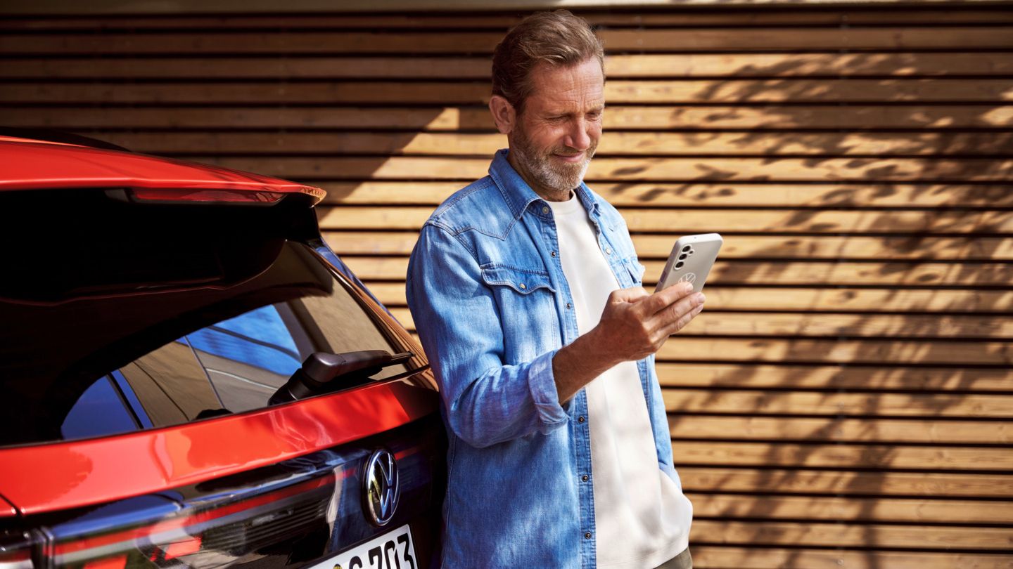 Ein Mann mit Smartphone in der Hand steht angelehnt an seinem Tiguan.