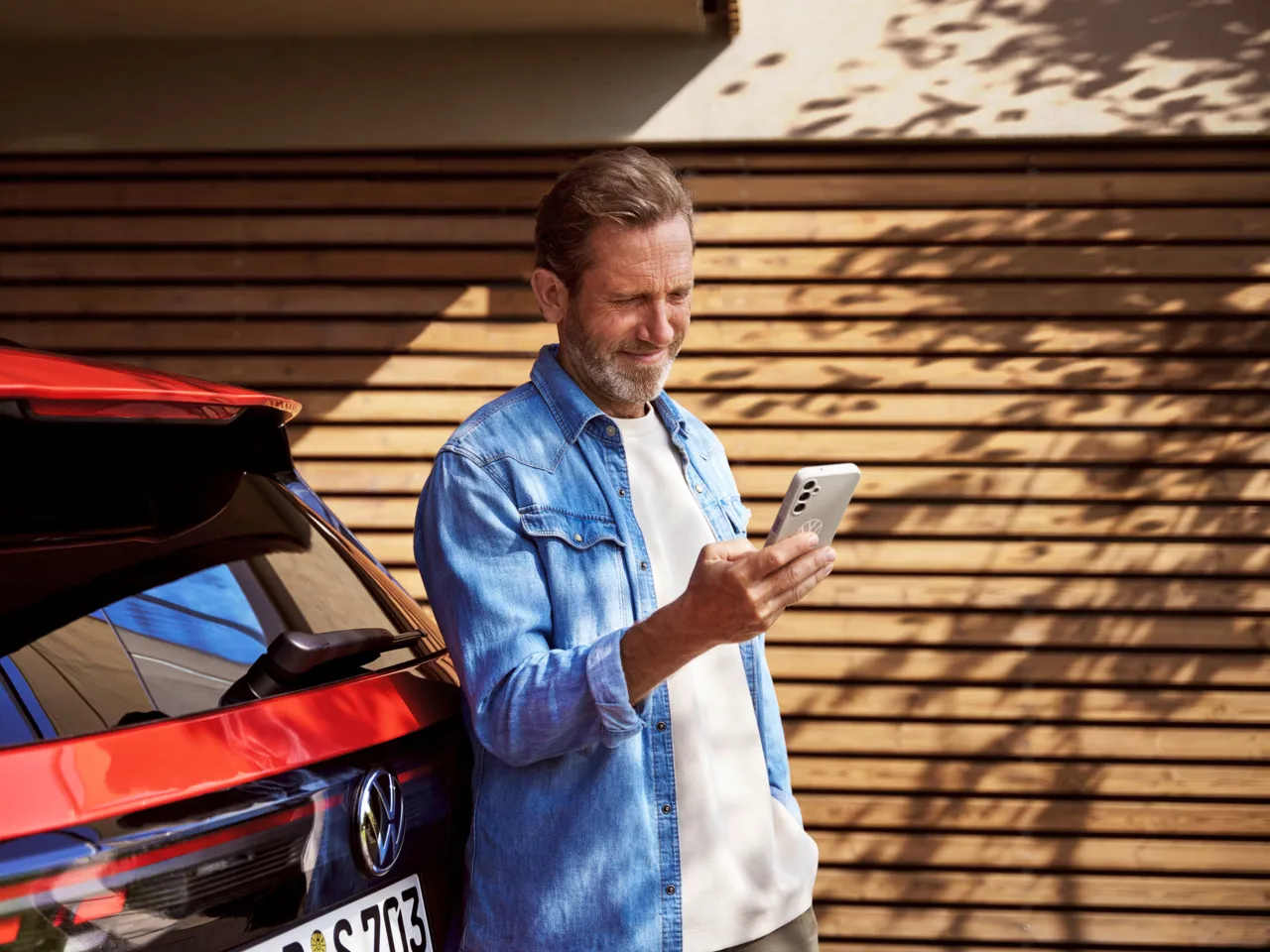 Ein Mann mit Smartphone in der Hand steht angelehnt an seinem Tiguan.