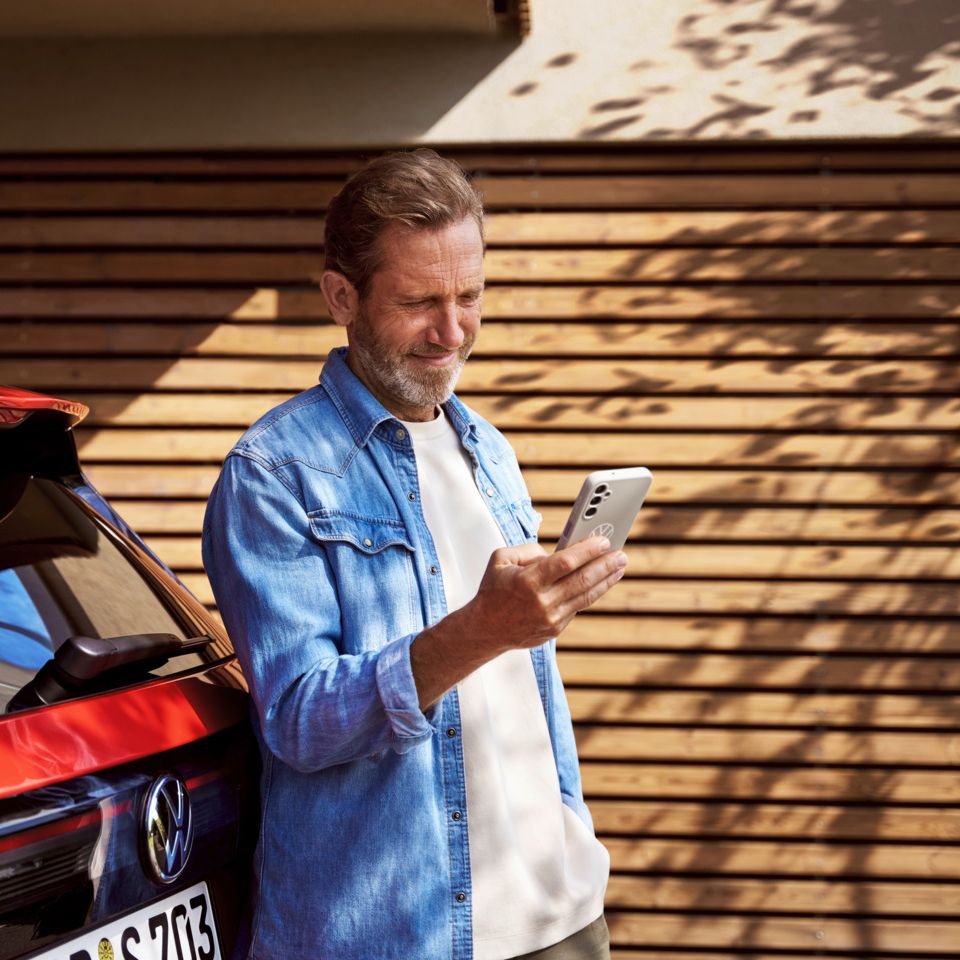 Ein Mann mit Smartphone in der Hand steht angelehnt an seinem Tiguan.