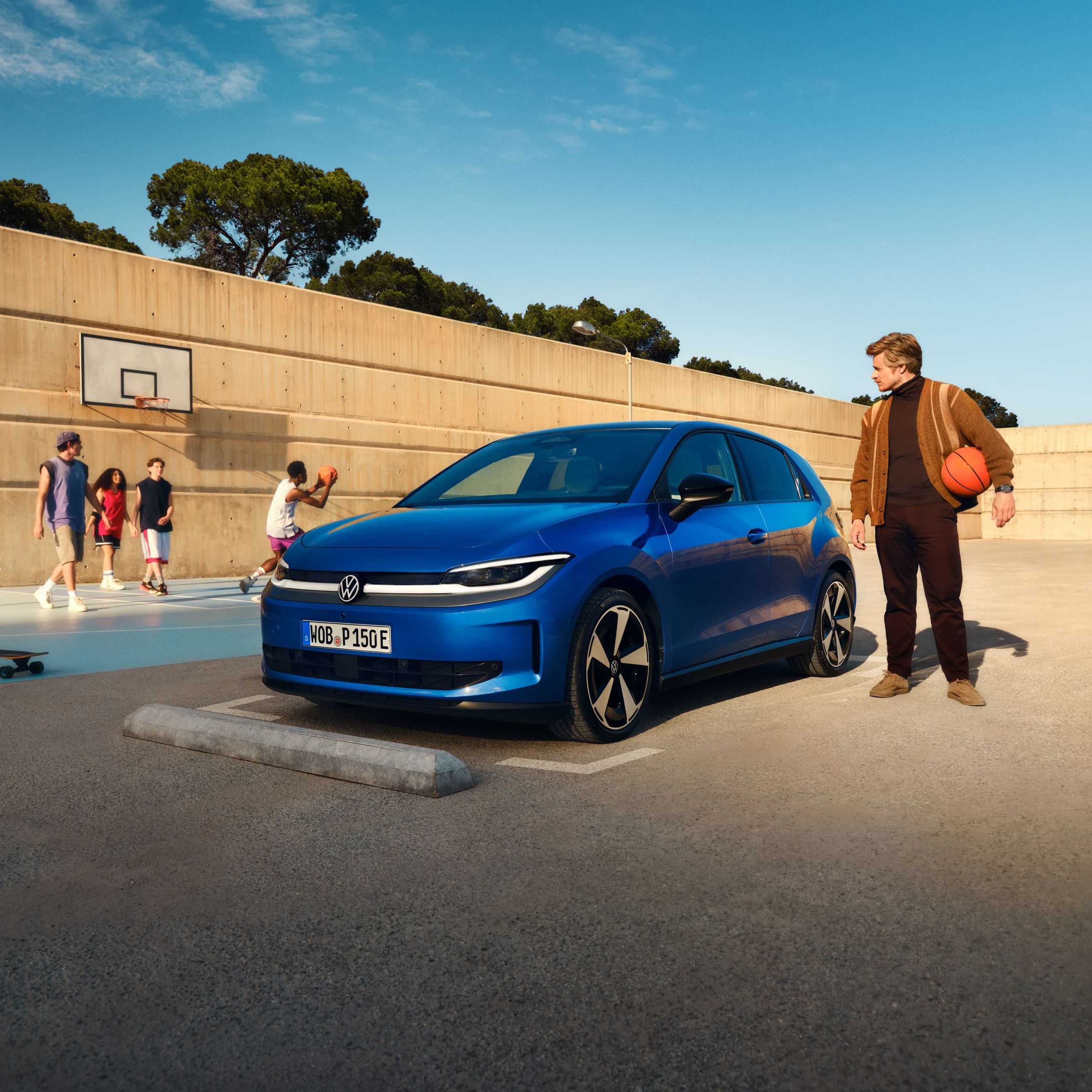 Um VW ID. Polo azul estacionado junto a um campo de basquete. Um homem está de pé nas proximidades, segura uma bola de basquetebol debaixo do braço.