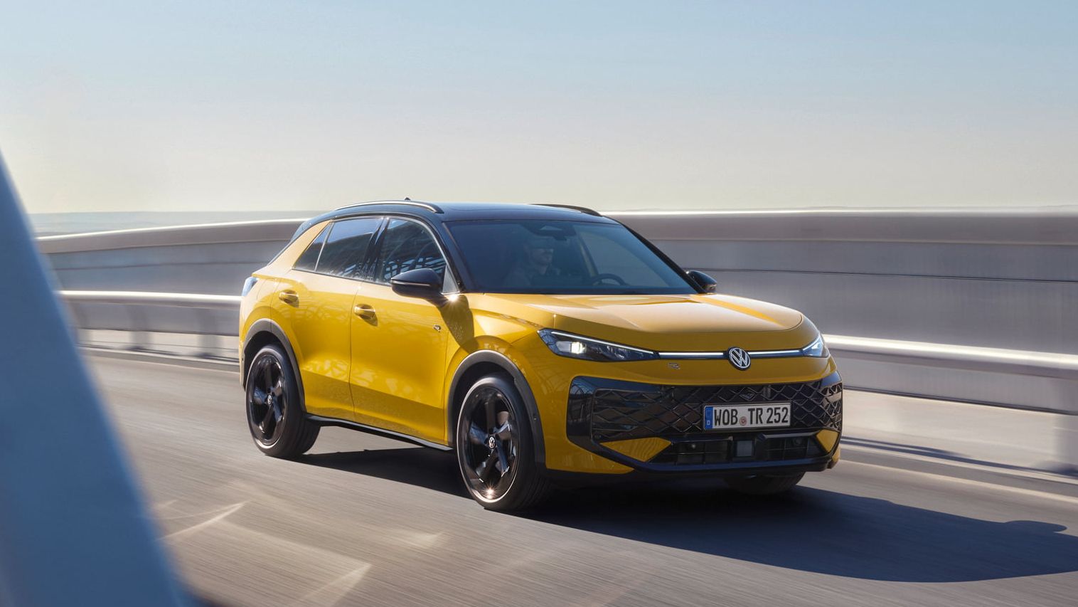Um VW T-Roc R-Line amarelo circula numa ponte com uma estrutura em aço moderna.