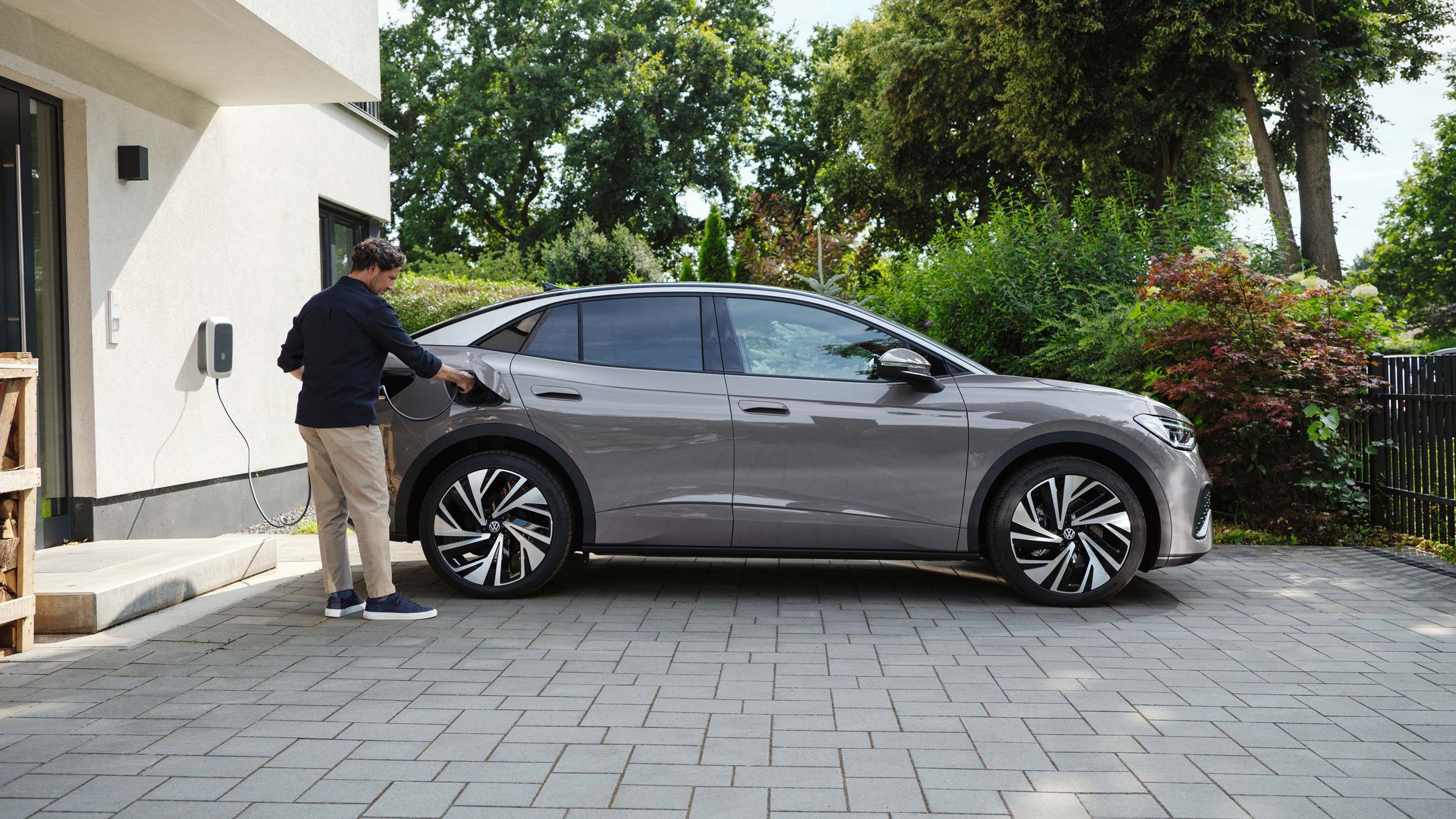 Homem parado ao lado do Volkswagen ID.5 em cor cinza, estacionado numa entrada de garagem pavimentada, carregando-o.