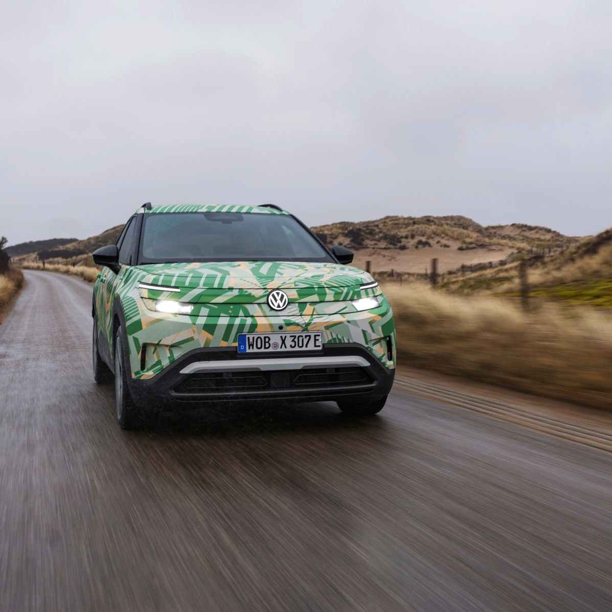 Ein Elektroauto der Marke VW fährt eine nasse Landstraße entlang. Die Landschaft rechts und links ist trocken und sandig, vermutlich eine Dünenlandschaft. Der Himmel ist bewölkt.