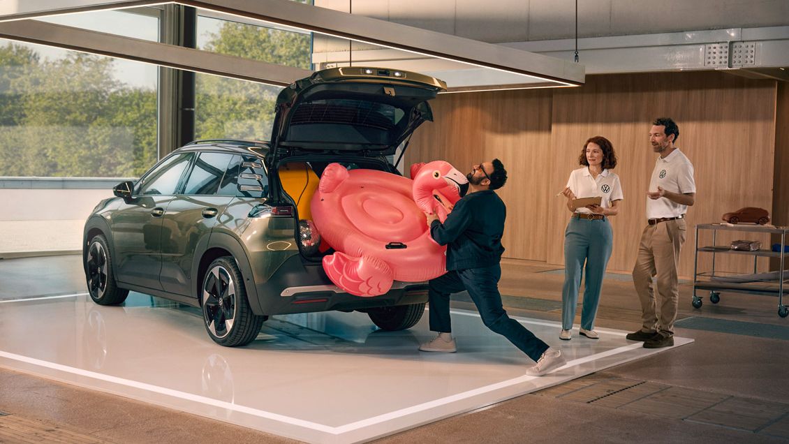 Um homem empurra um flamingo insuflável para dentro da bagageira aberta do VW ID. Cross. Duas pessoas estão ao seu lado e divertem-se a olhar para ele.