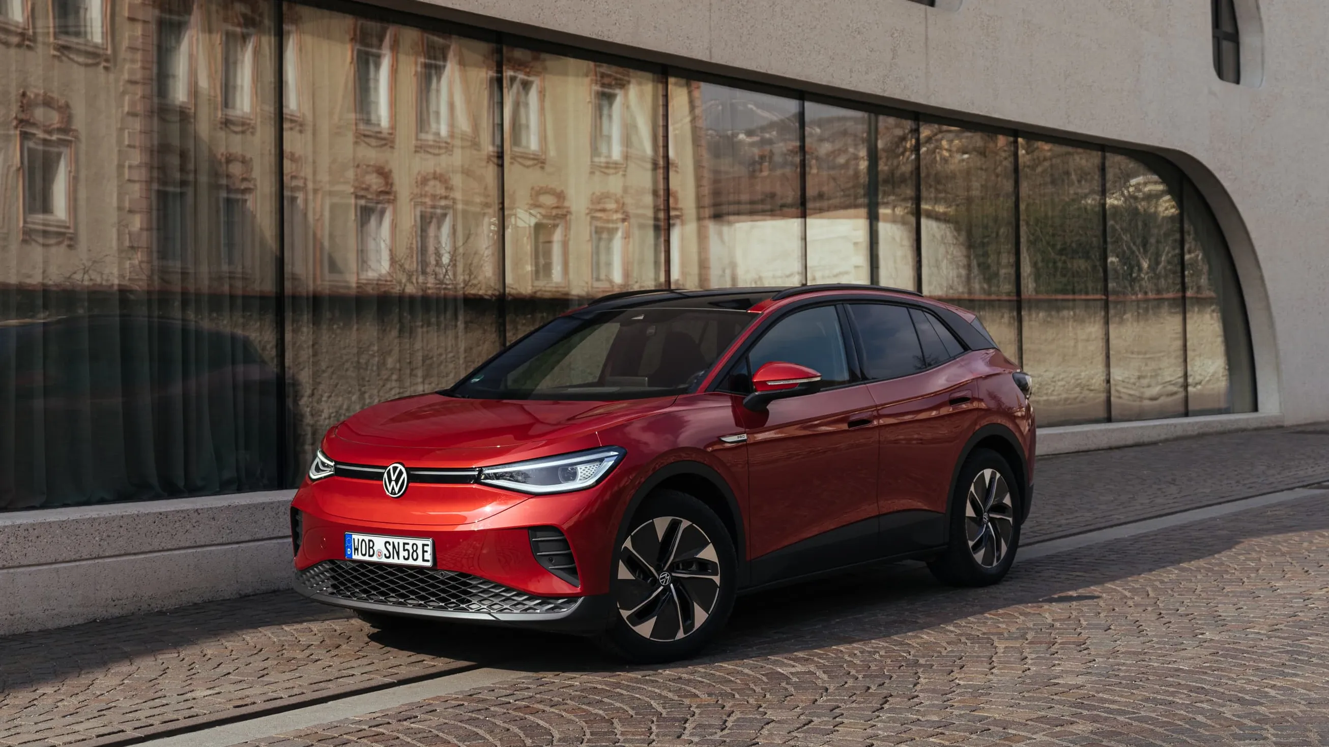 Veículo elétrico Volkswagen ID.4 de cor vermelha estacionado em frente a um edifício moderno com janelas altas e estreitas.