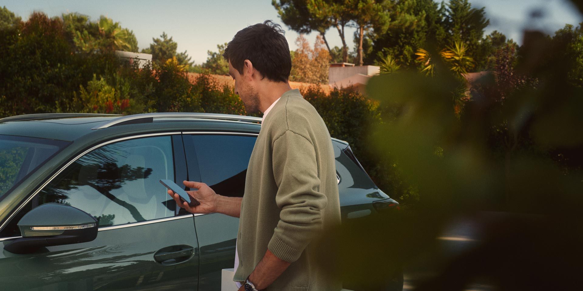 Um jovem no lado do condutor do VW Tiguan verde escuro opera o Park Assist Plus opcional através do seu smartphone.